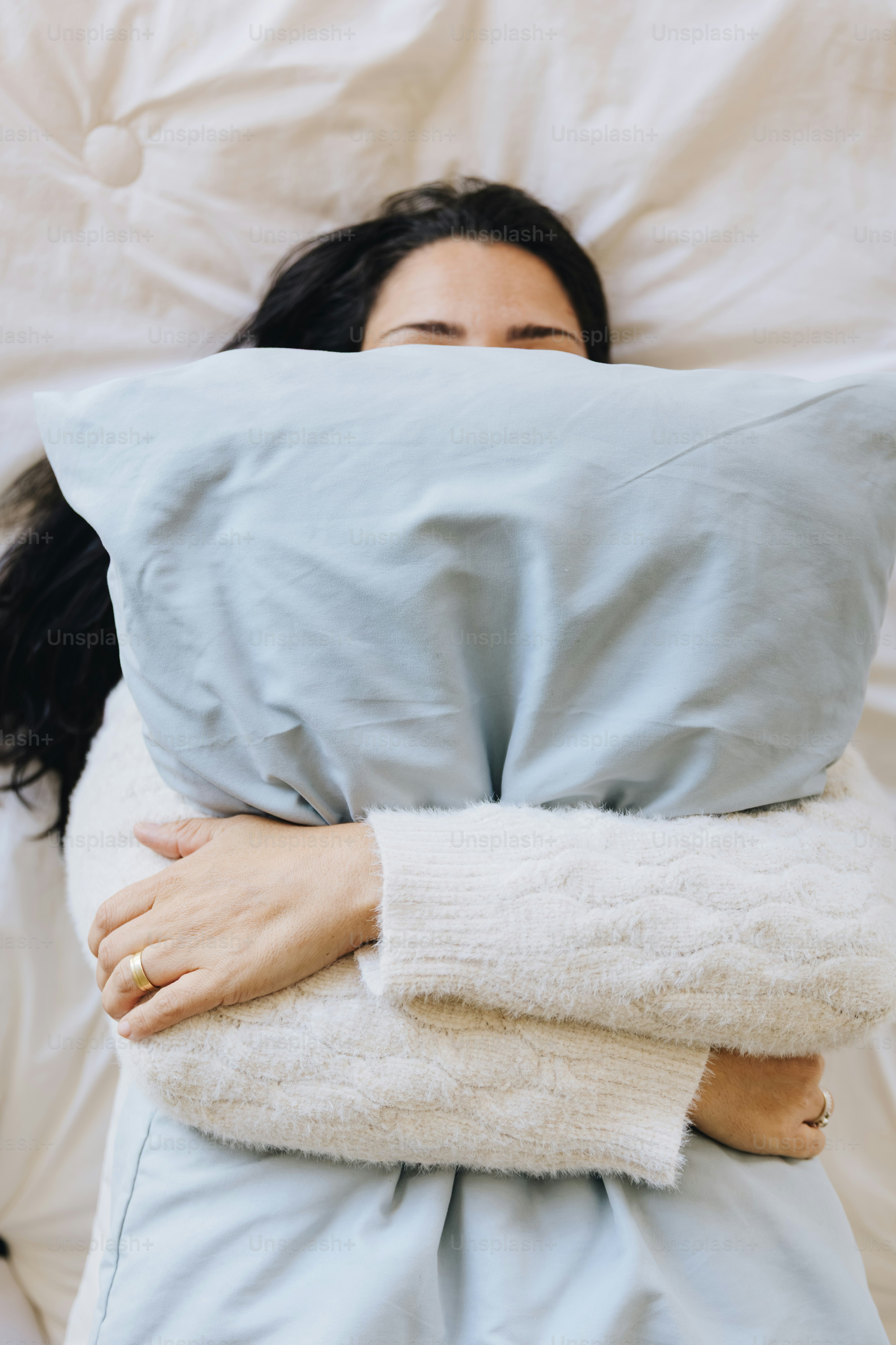 A person in a white sweater hugging a light blue pillow in bed