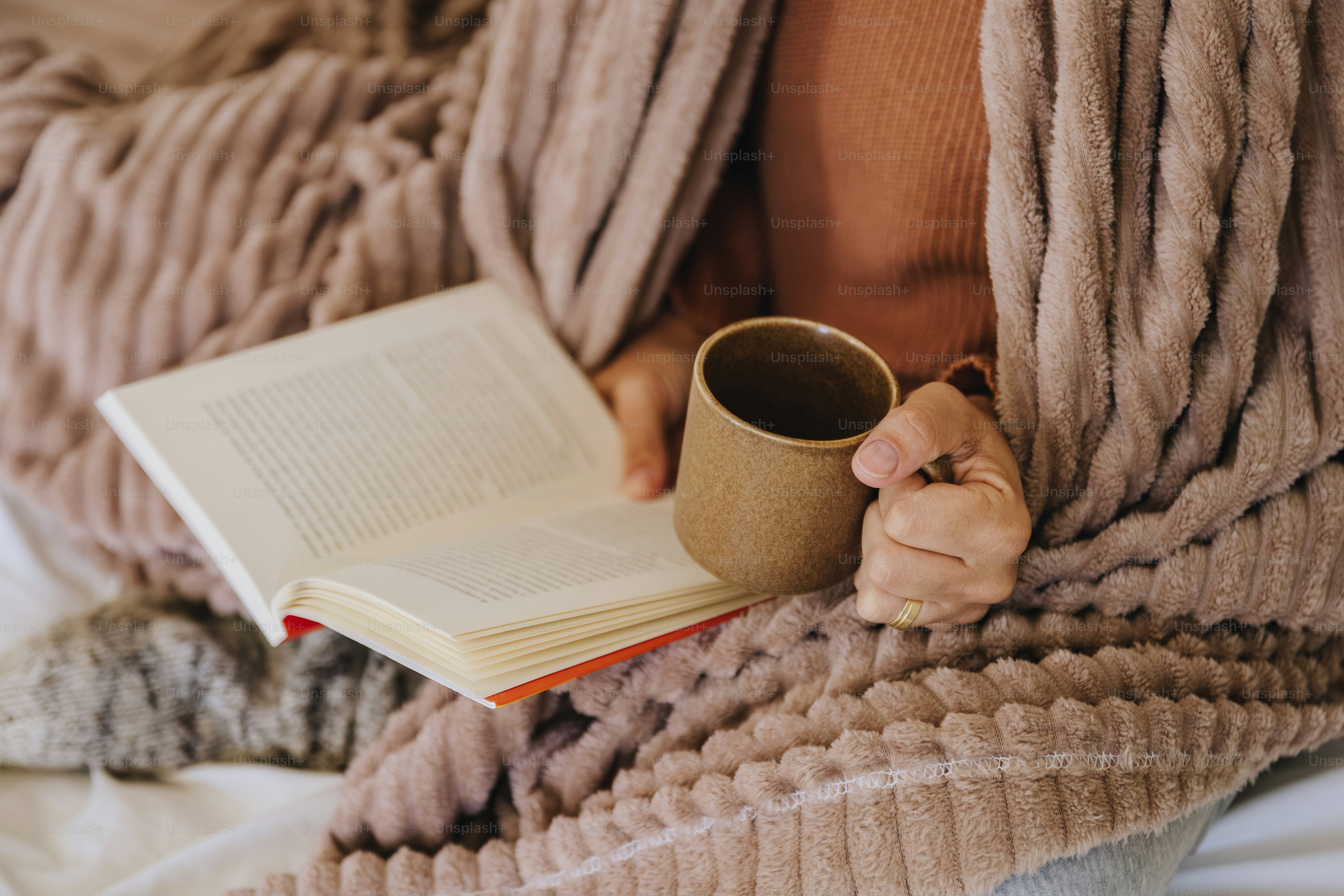 A person holding a book and a brown mug while wrapped in a blanket