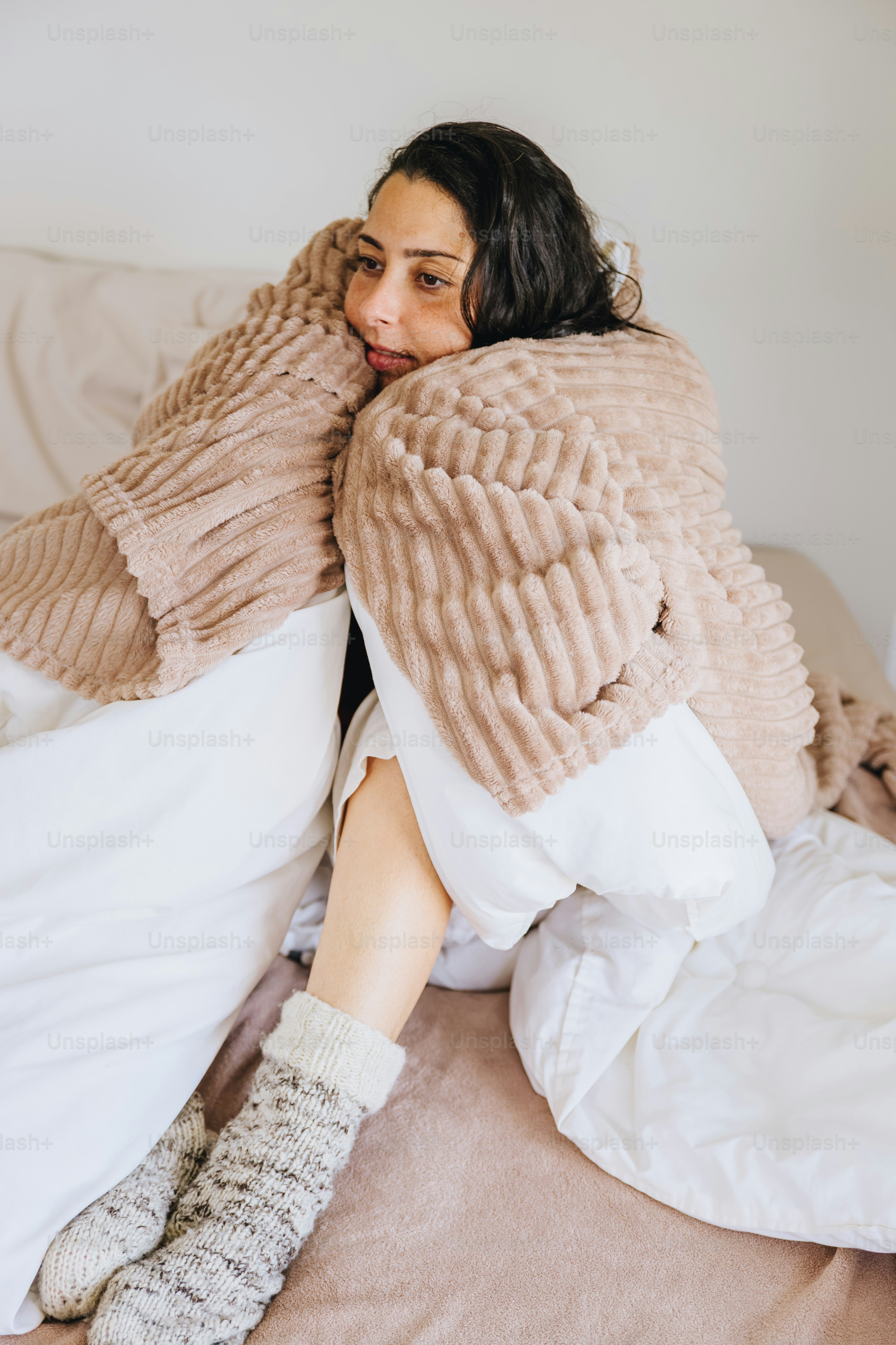 A woman wrapped in a textured beige blanket wearing wool socks on a bed