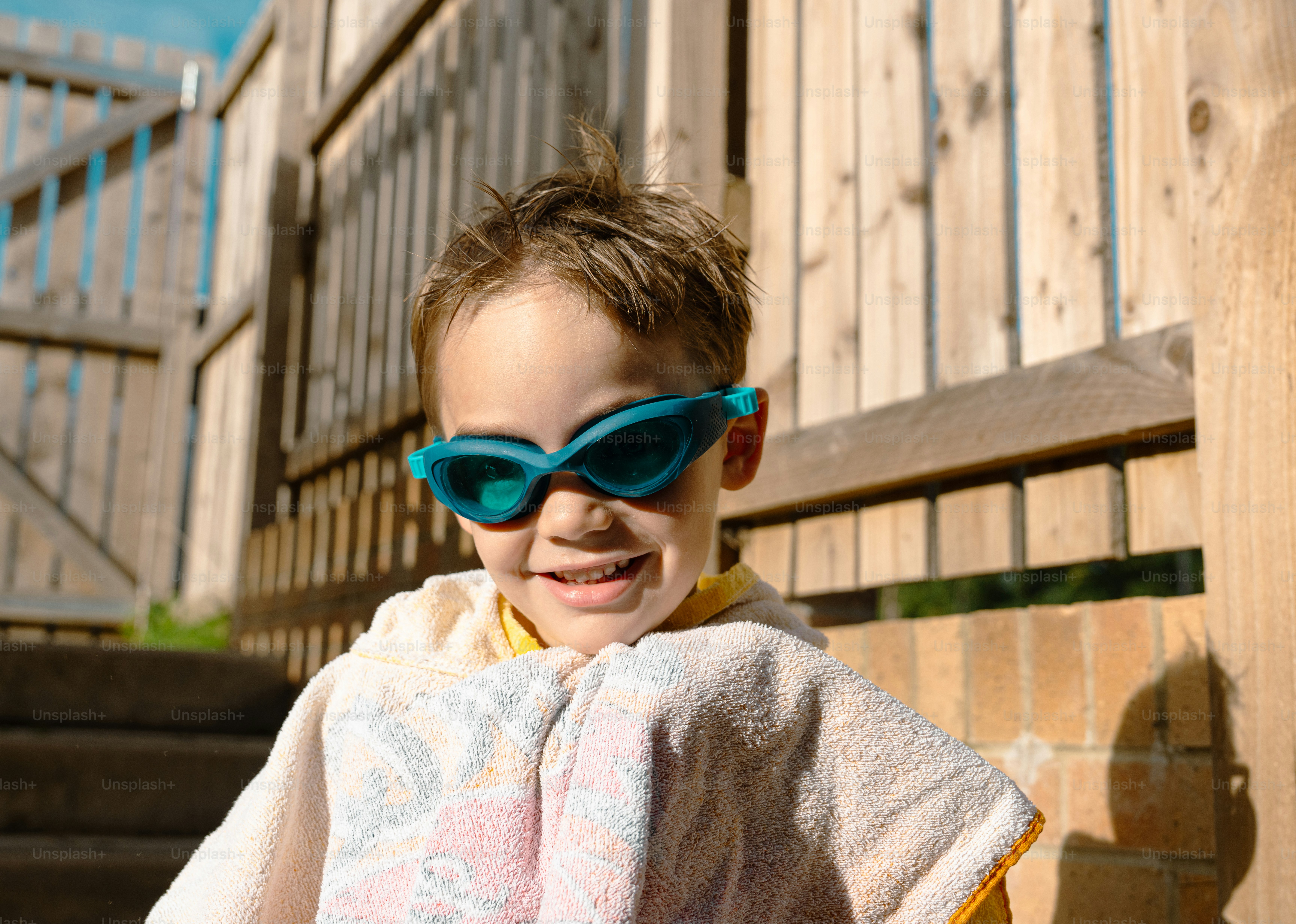 A smiling child wearing teal swimming goggles and a towel in a sunlit yard