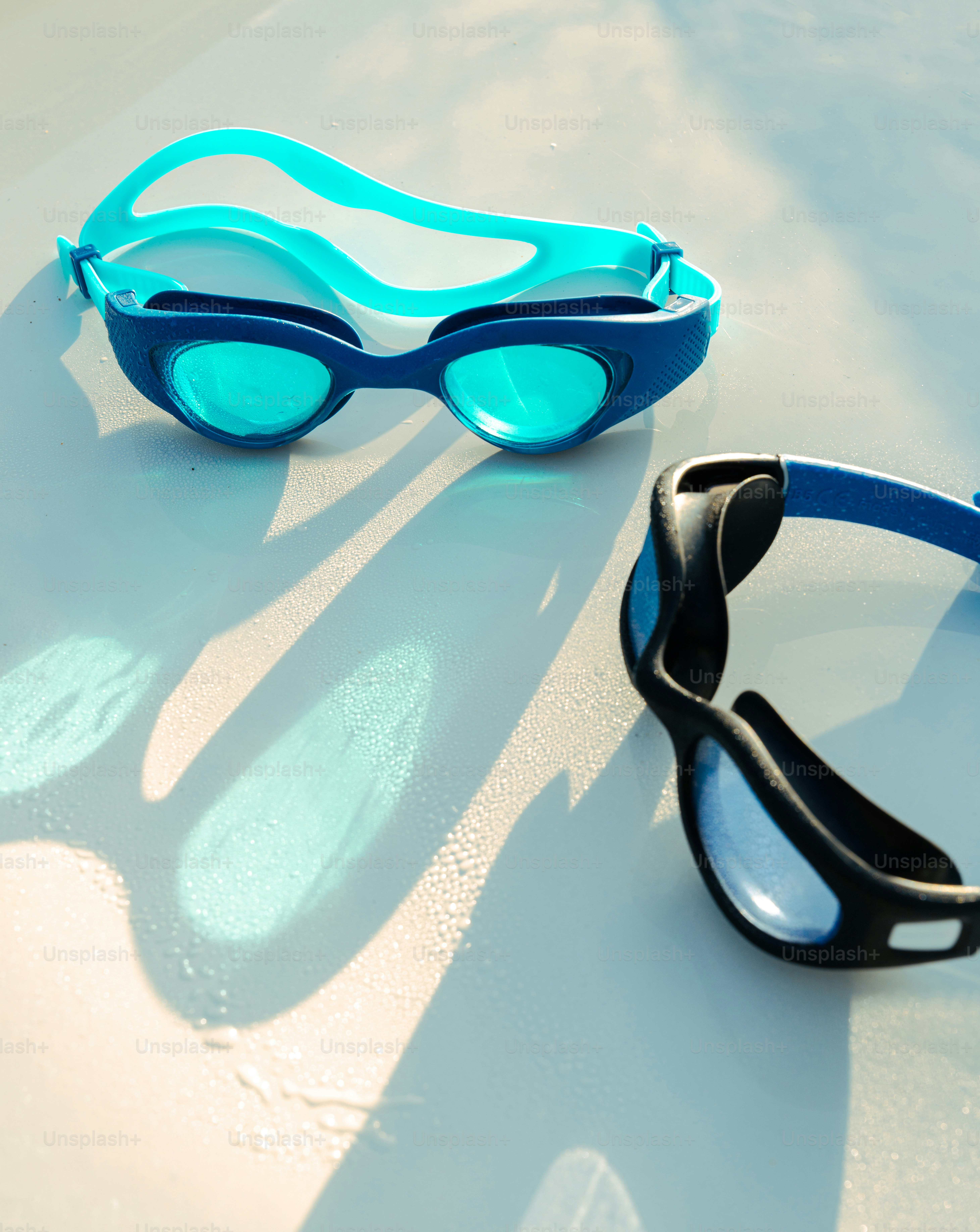 Two pairs of swimming goggles on a wet white surface in bright sunlight