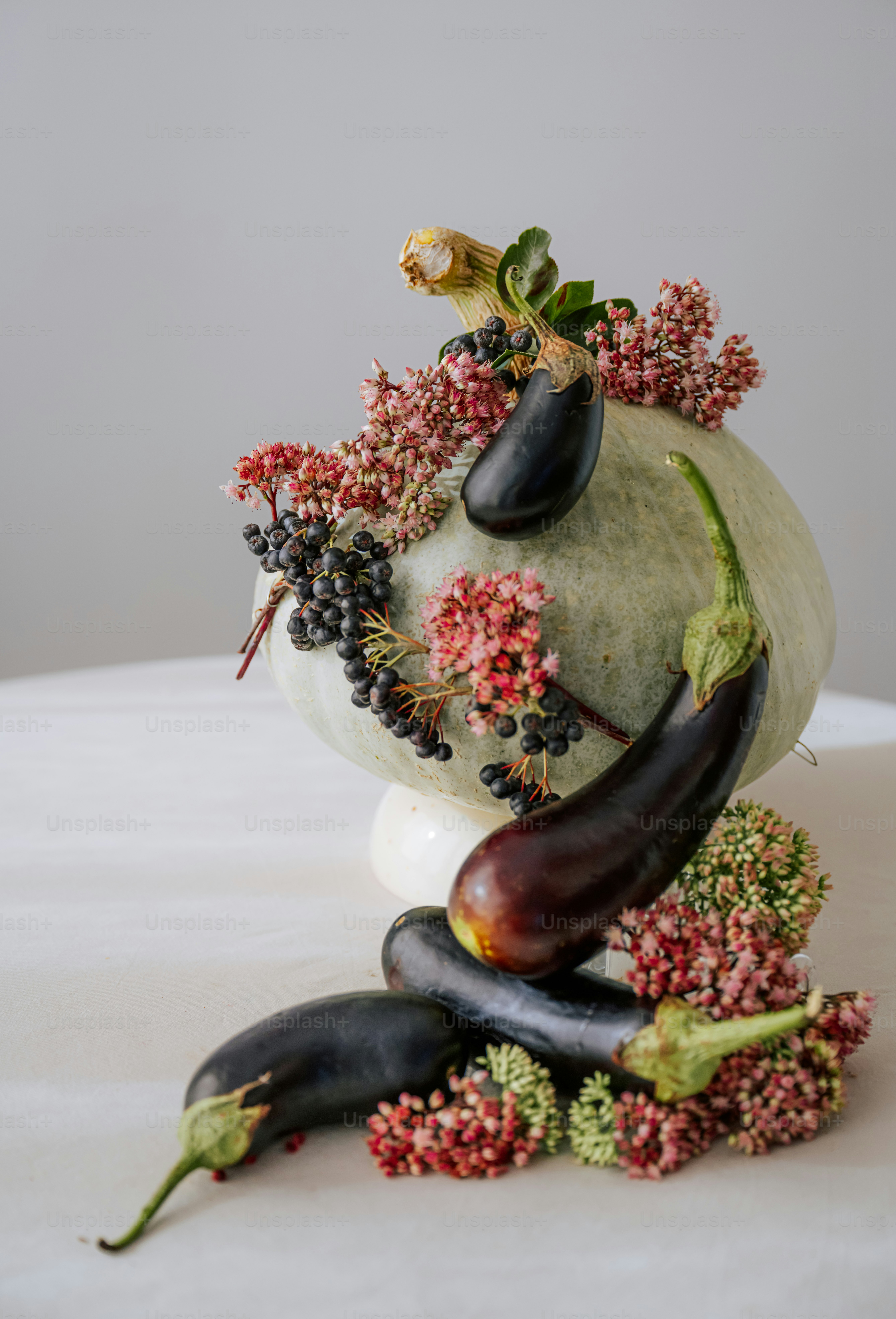 A pale green pumpkin decorated with dark eggplants, black berries, and pink flowers