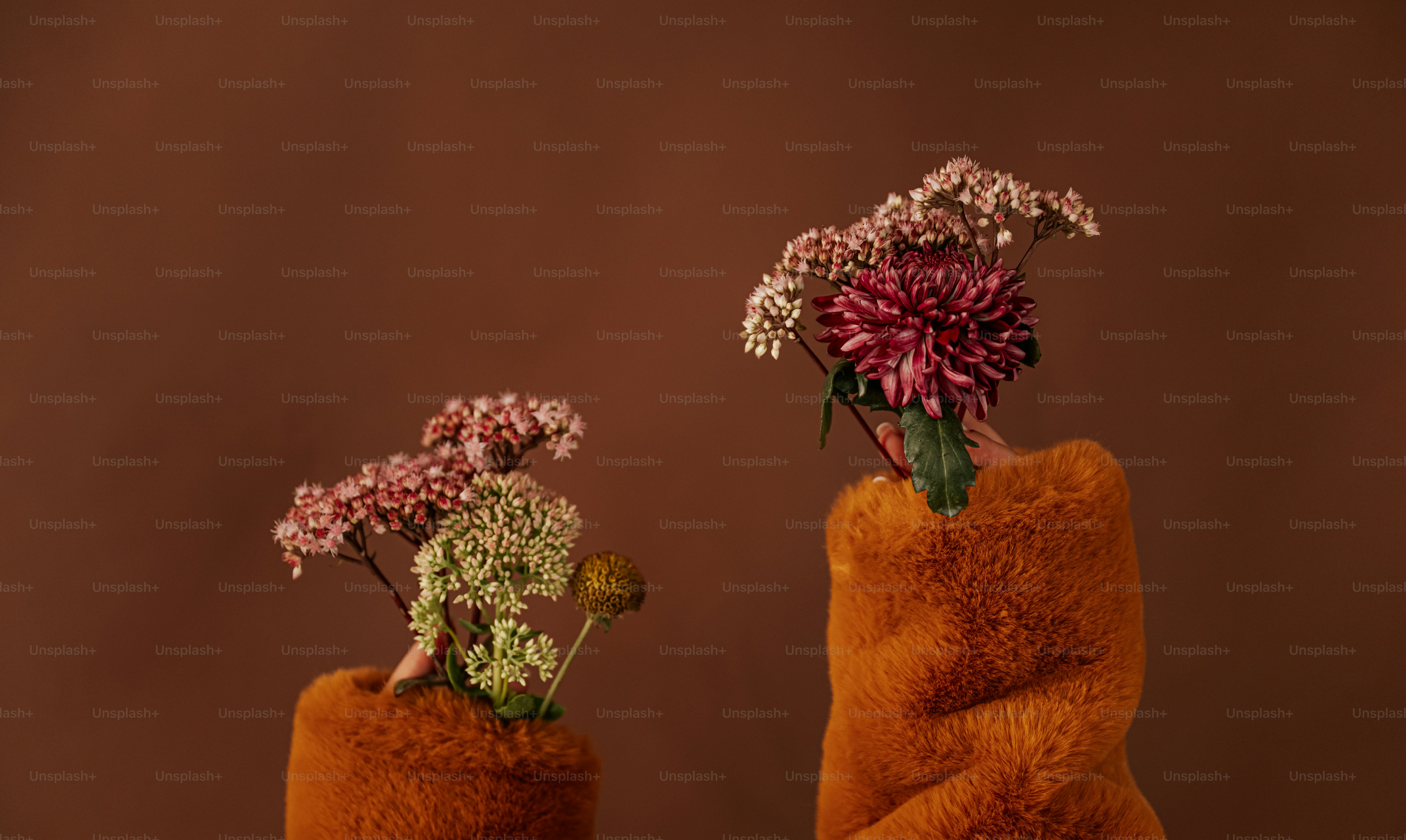 Two hands in fuzzy orange sleeves holding small bouquets of flowers against brown