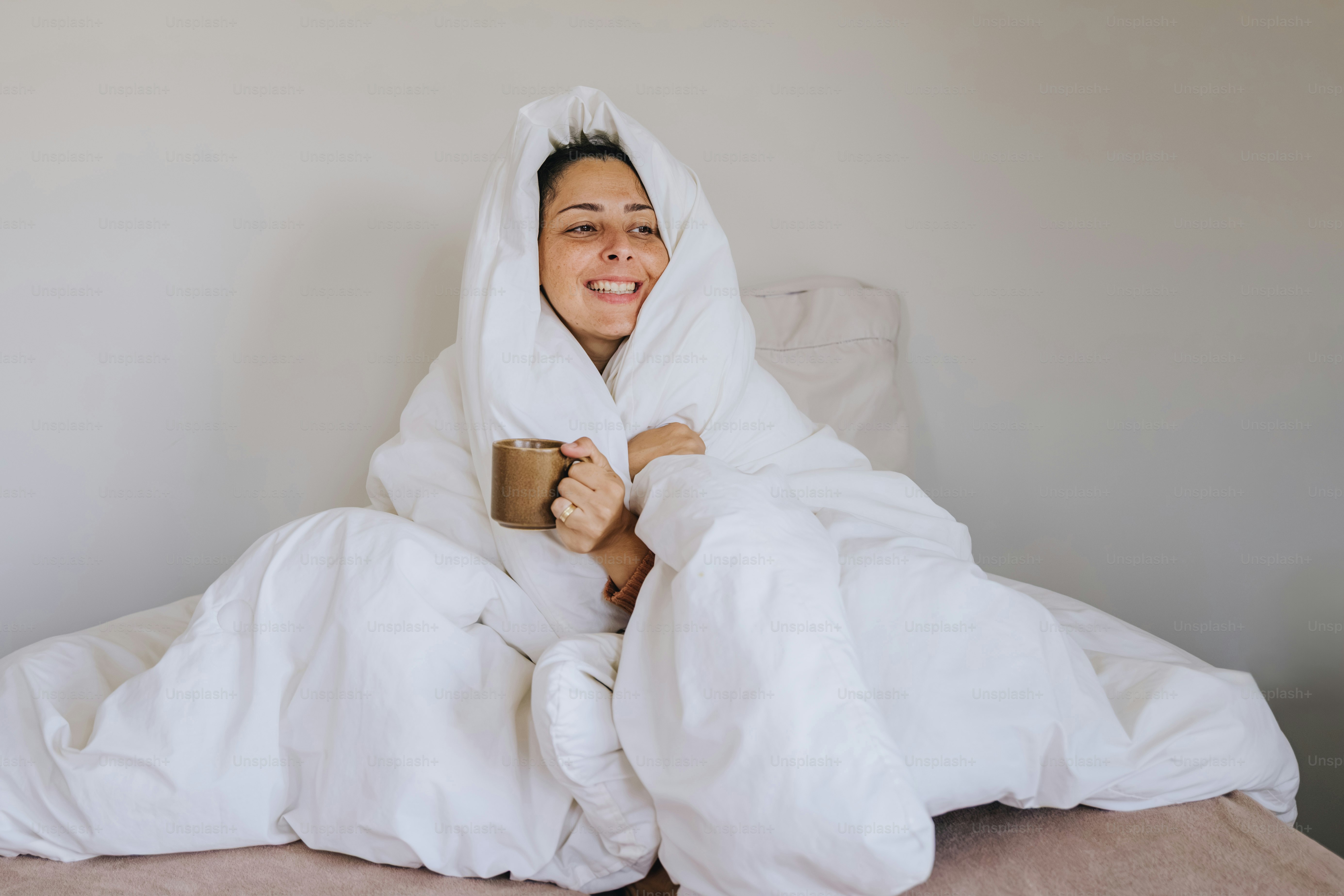 A smiling woman wrapped in a white duvet holding a brown ceramic mug