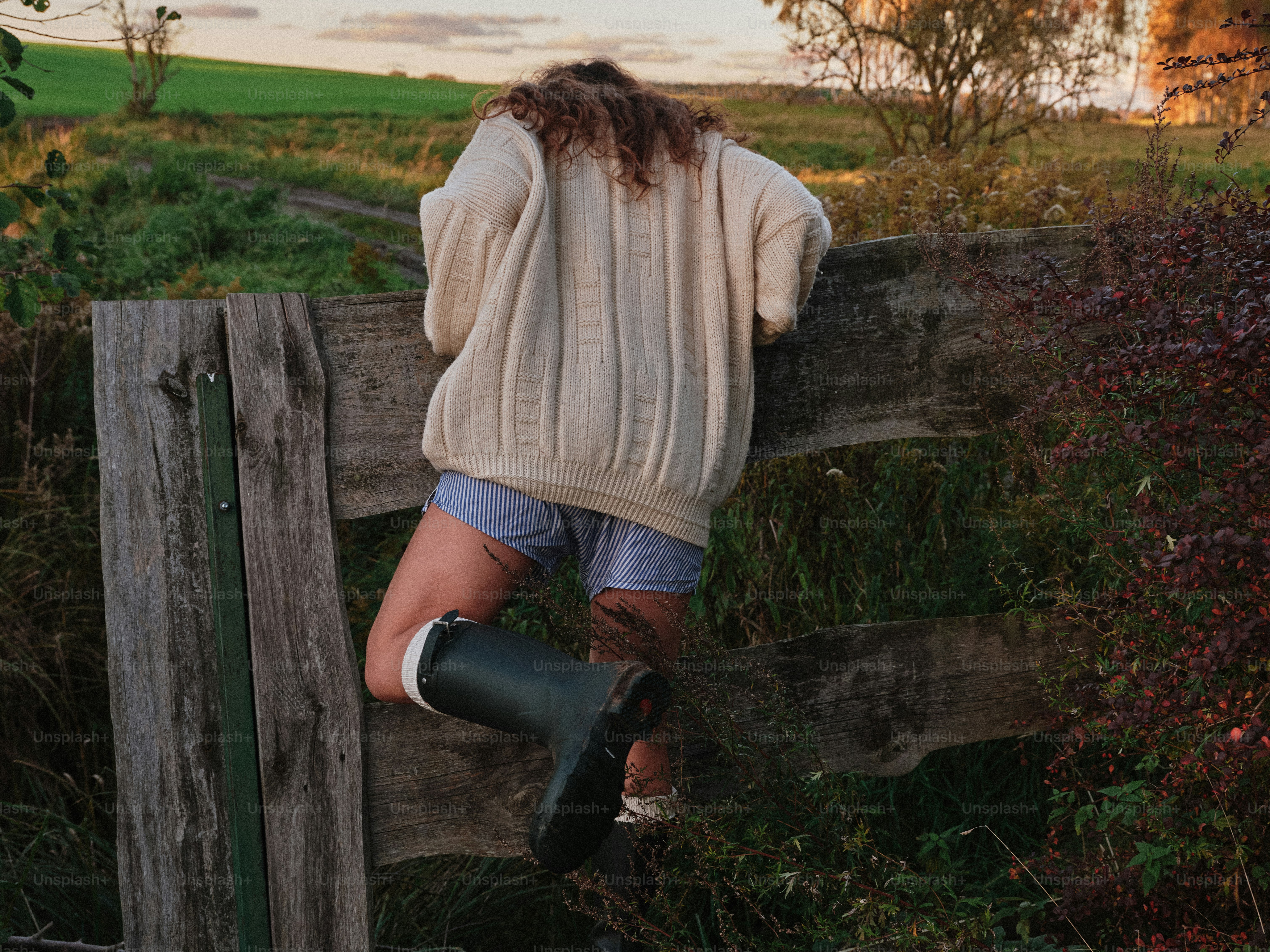 A person in a cream sweater climbing over a wooden fence in a field