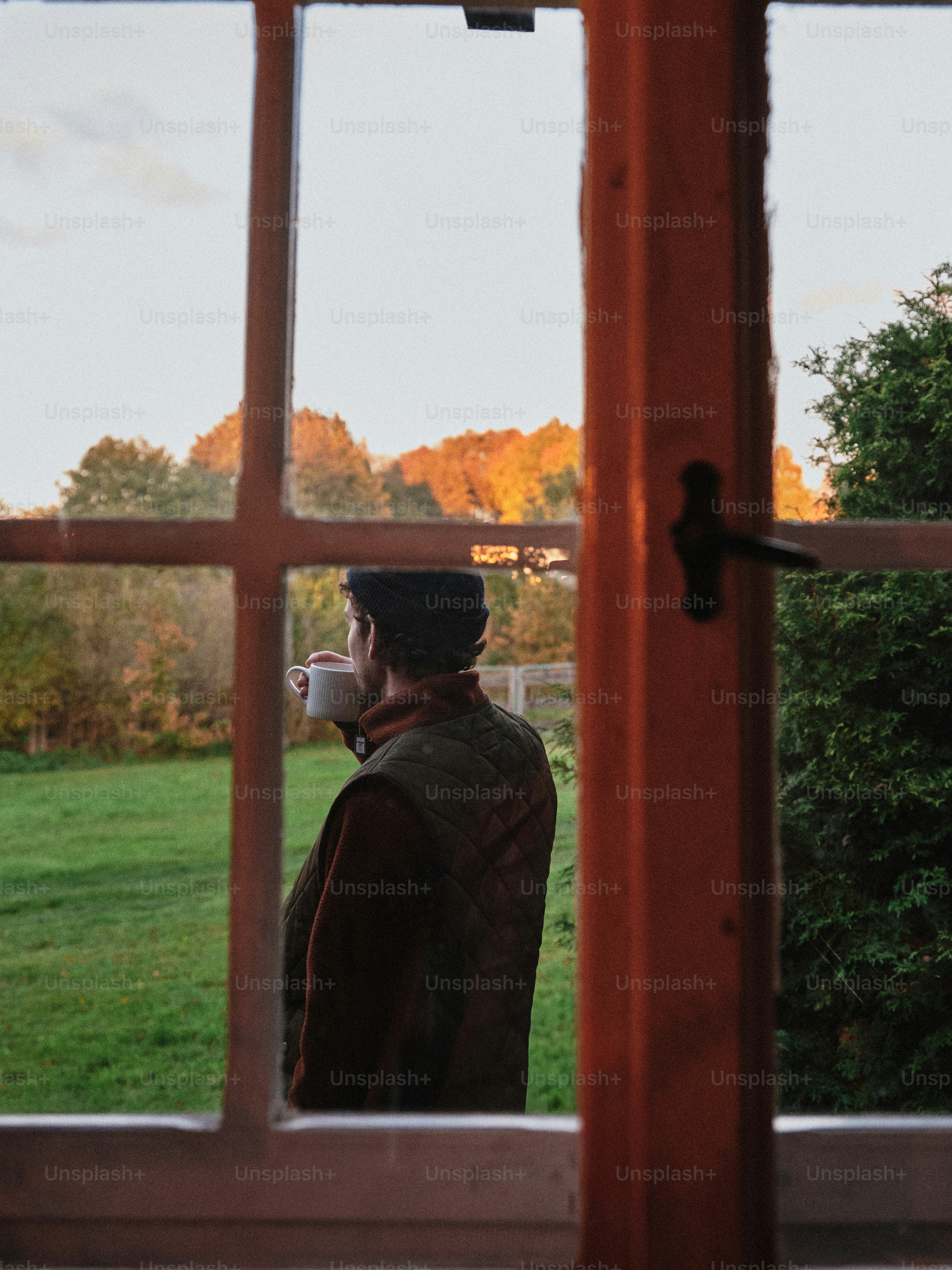 A person in a beanie and quilted vest drinking from a mug by a window