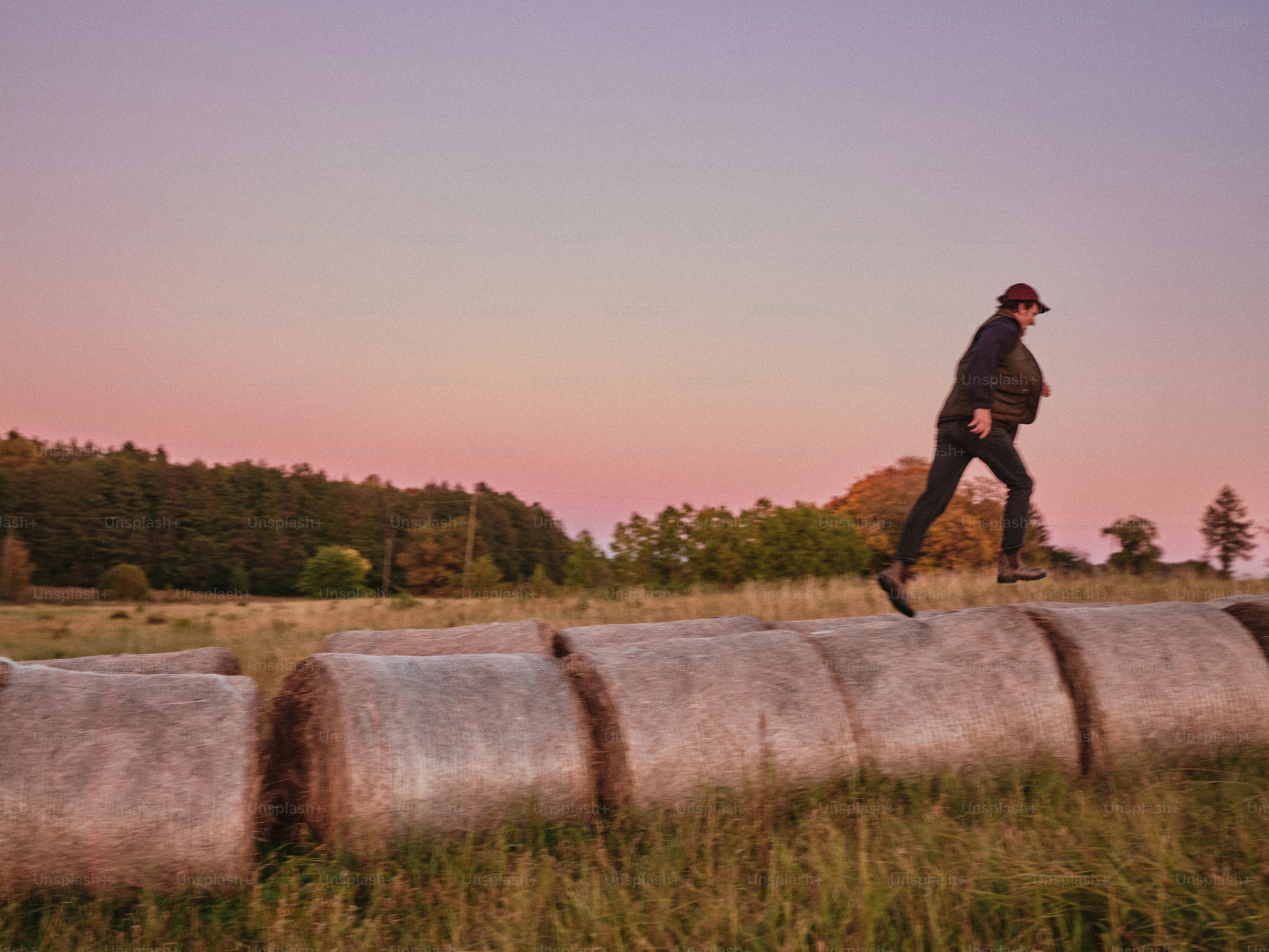 A person in a vest and hat jumping over hay bales in a field