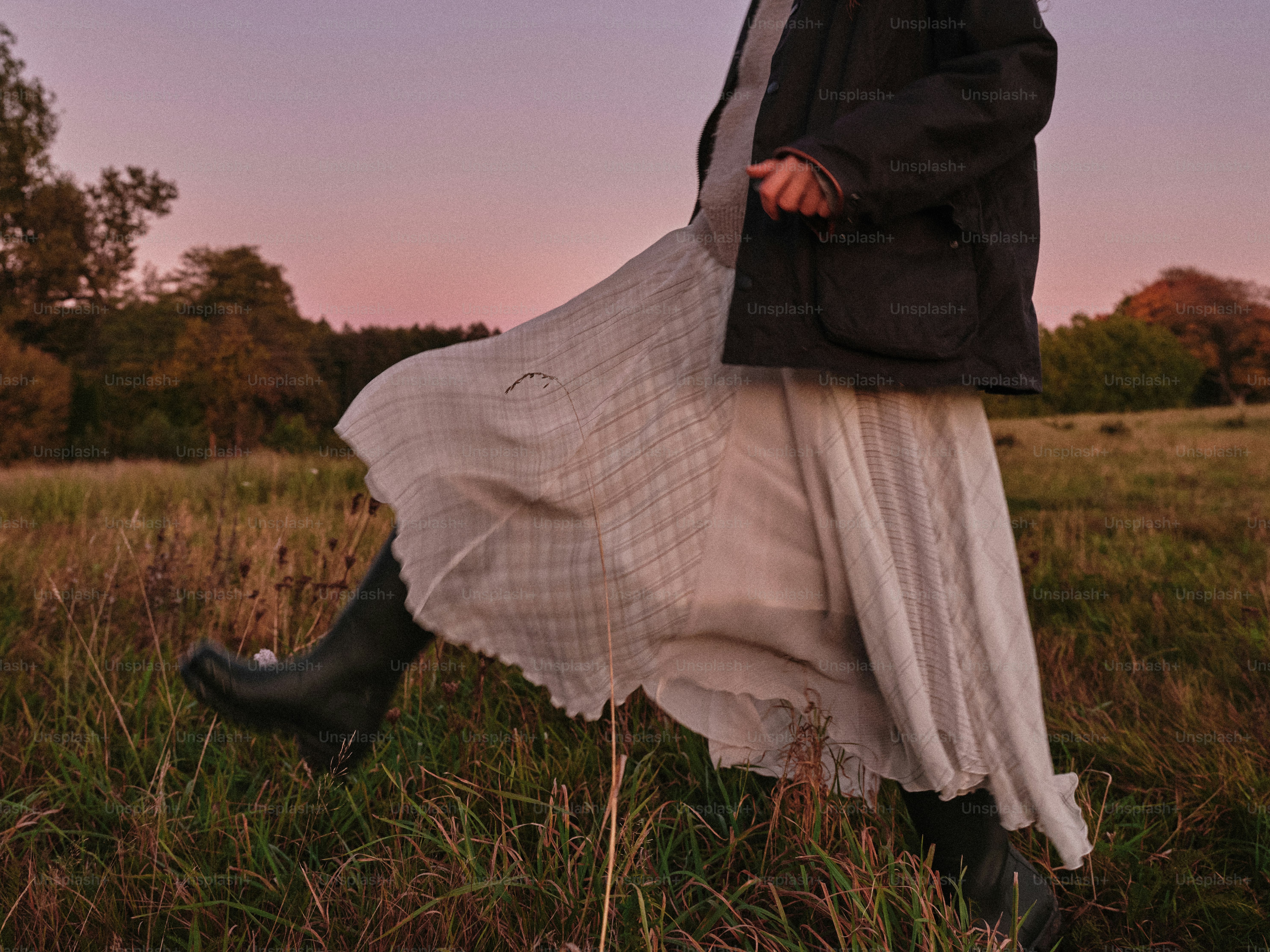 A person in a long skirt and dark jacket walking through a grassy field