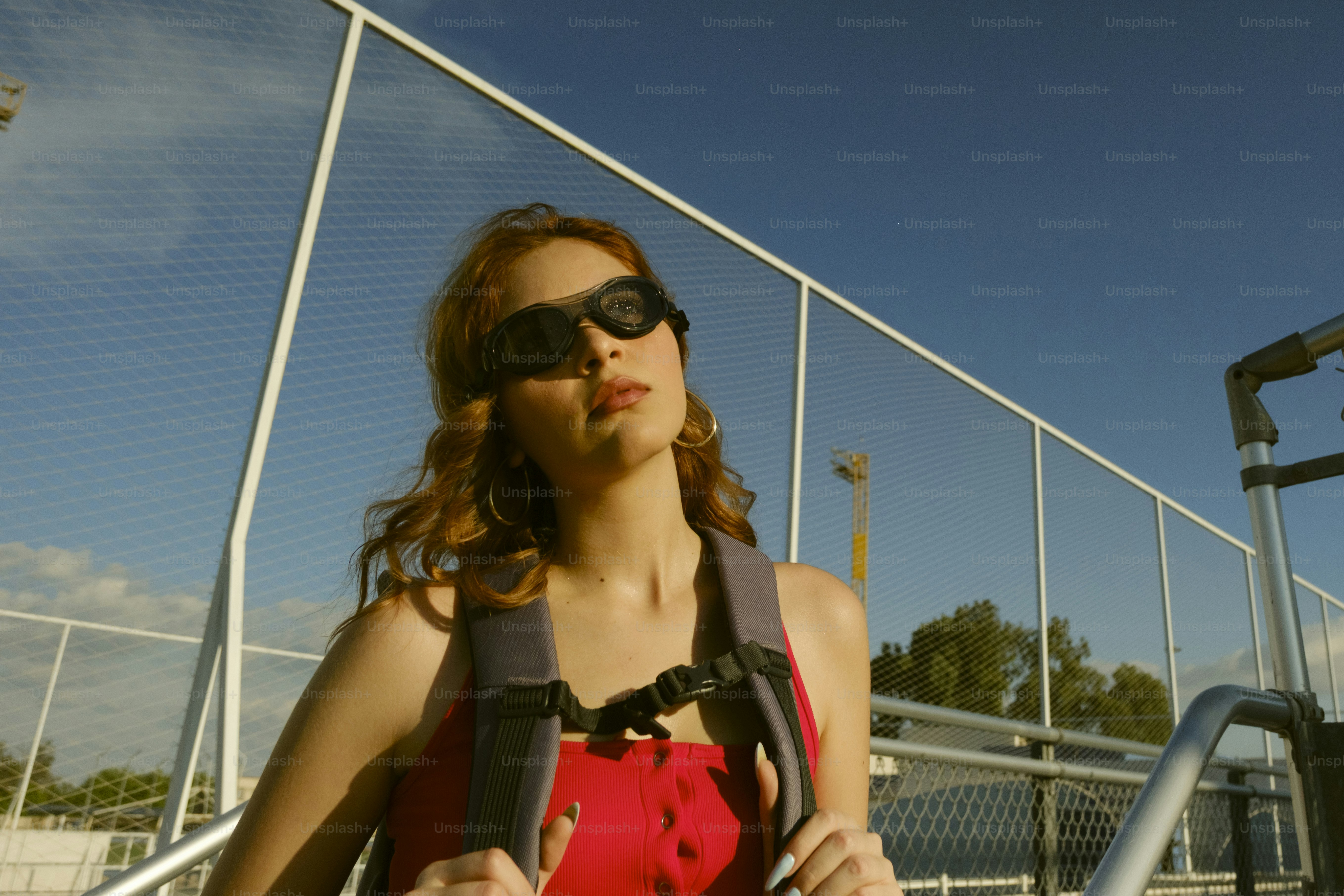 A woman with red hair wearing goggles and a backpack against a chain-link fence