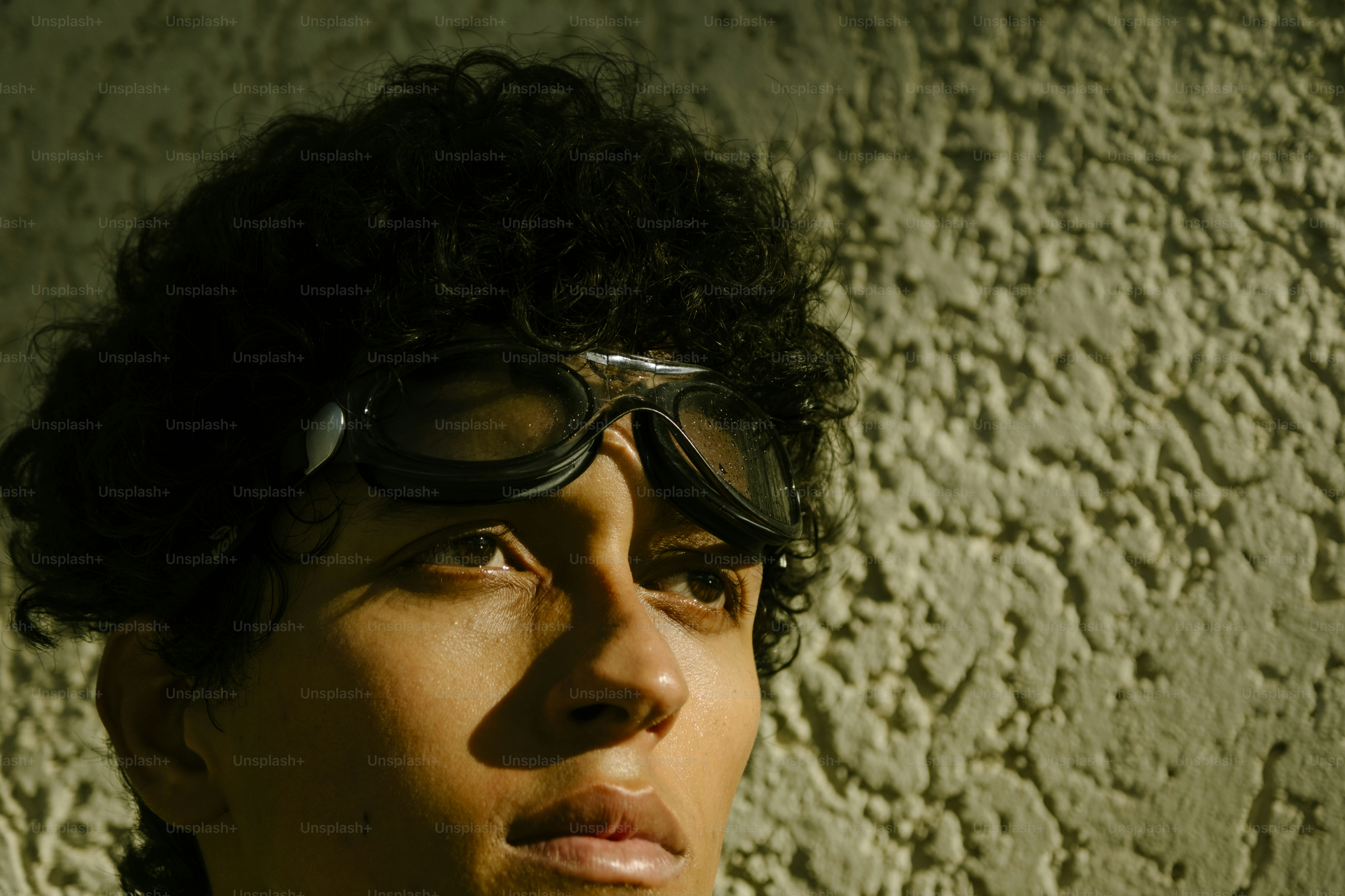 A person with curly hair wearing dark goggles against a textured wall