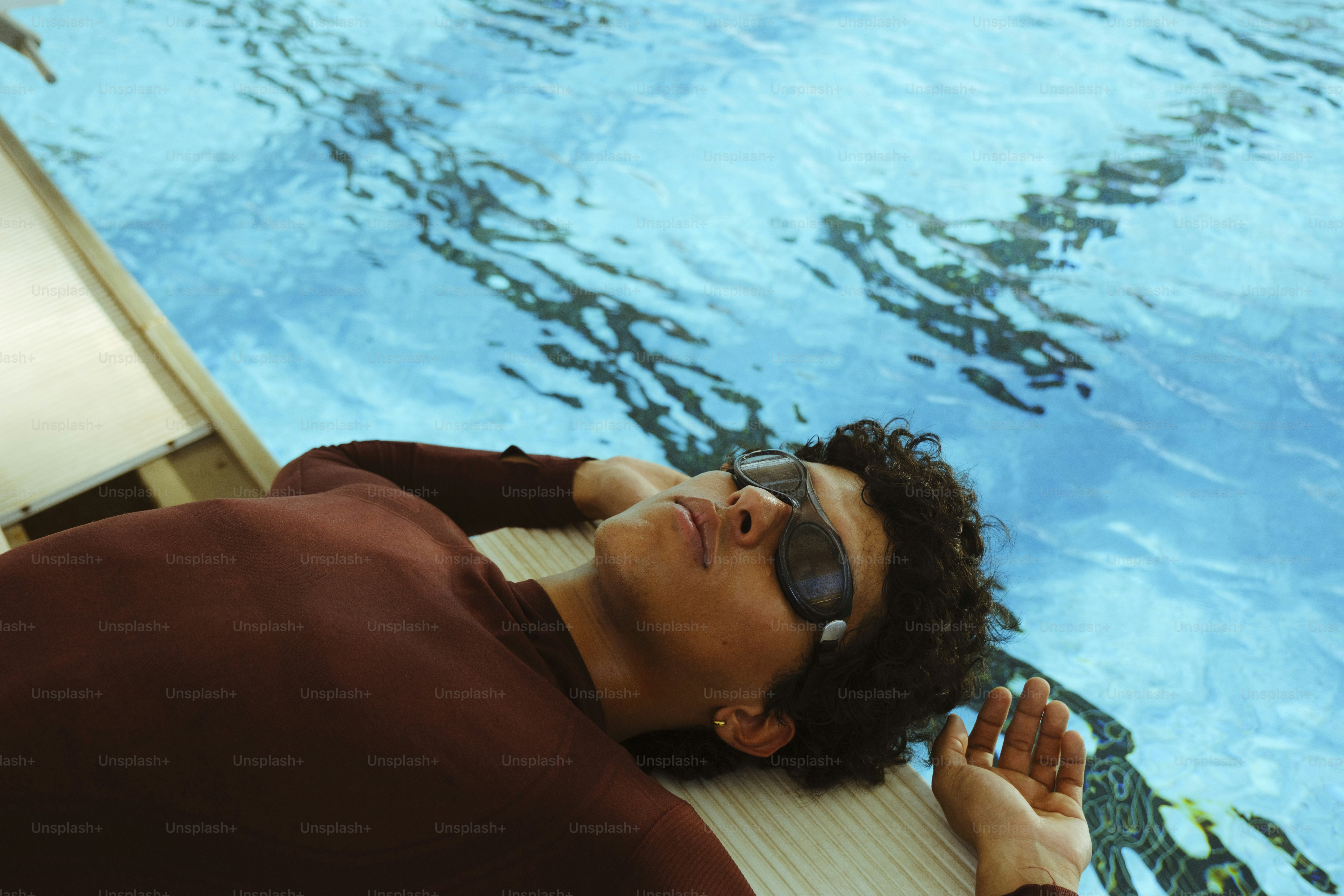 Uma pessoa com camisa marrom de manga longa e óculos de natação descansando à beira de uma piscina
