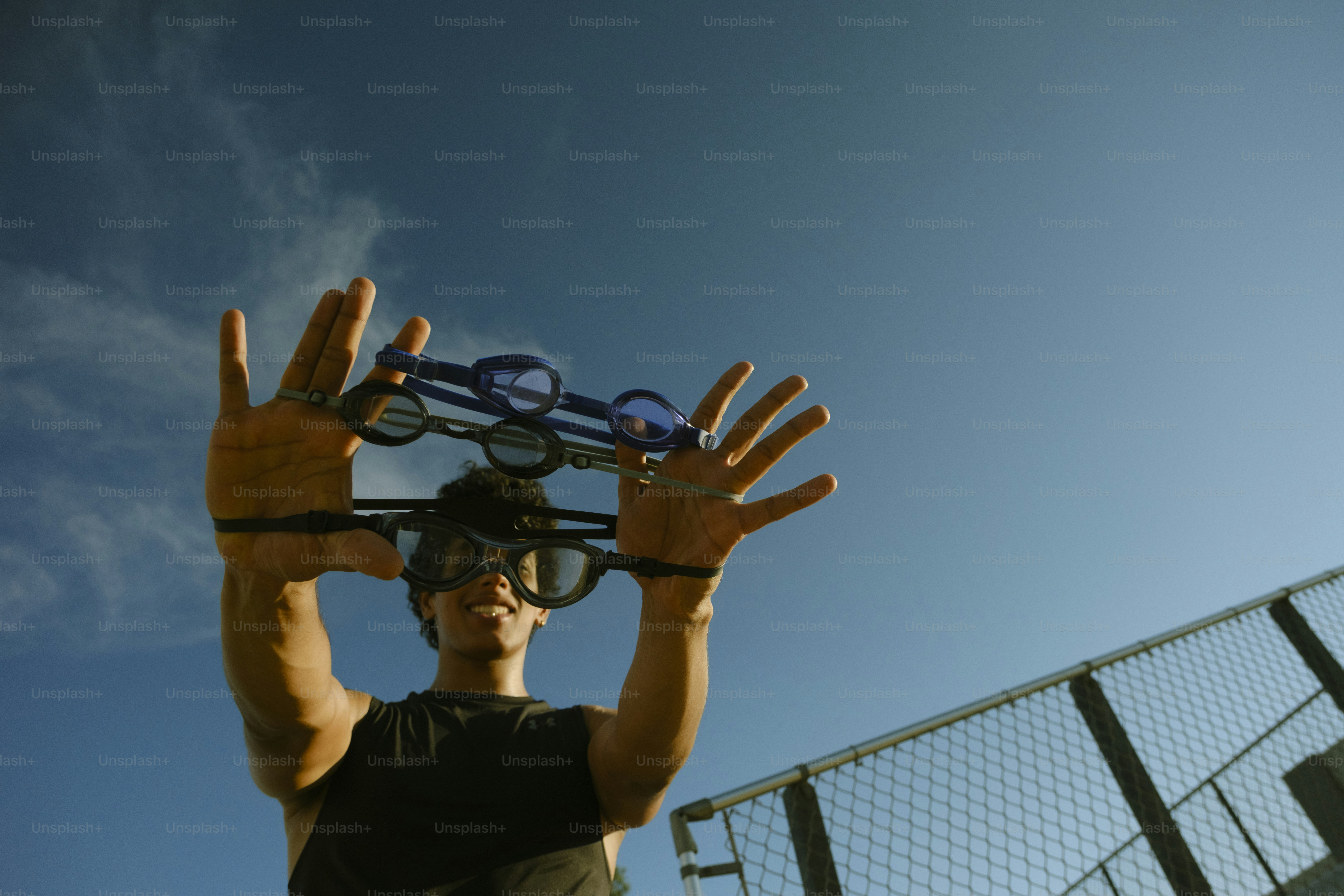 A person holding three pairs of swimming goggles against a clear blue sky
