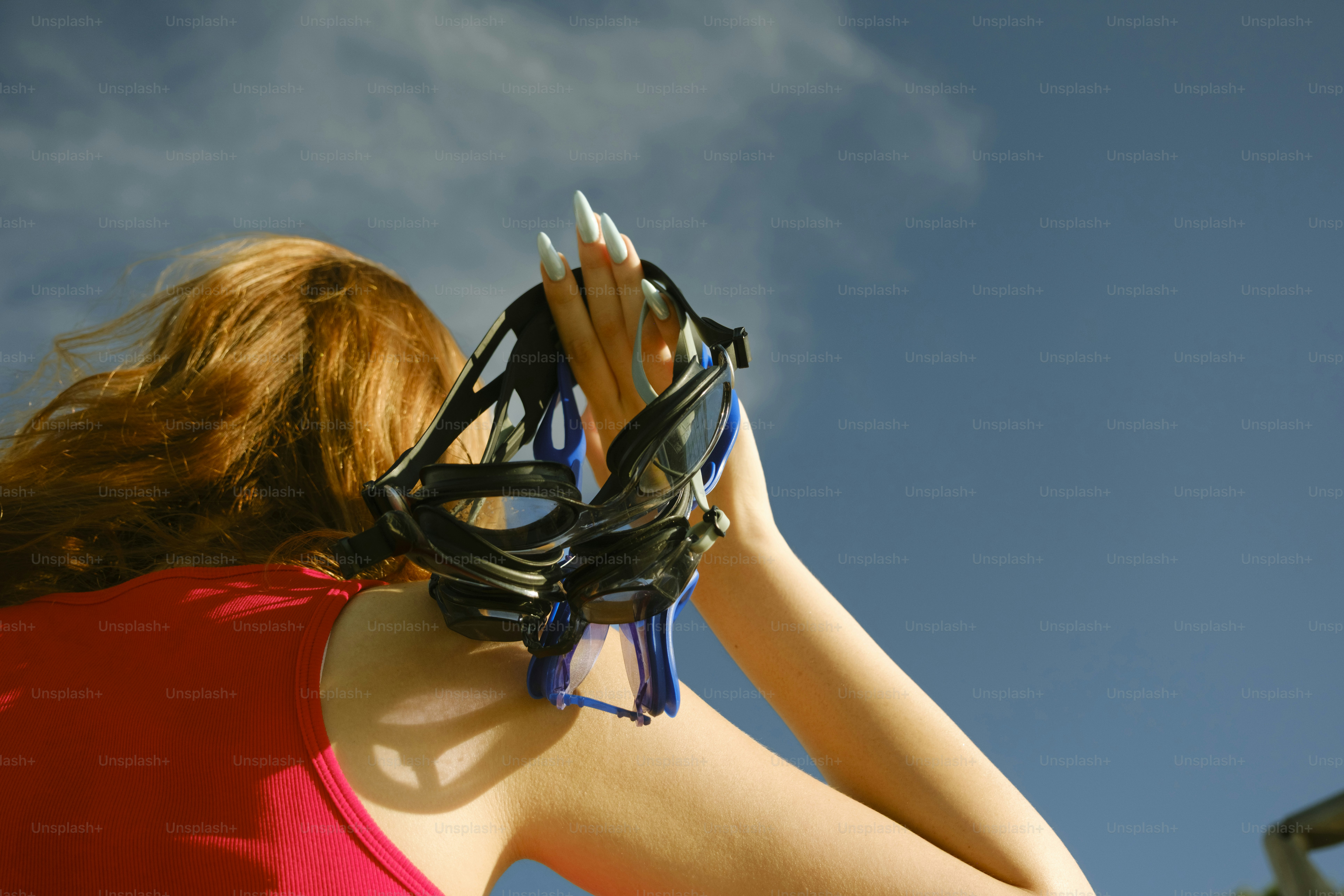 A person in a red tank top holding swimming goggles against a blue sky