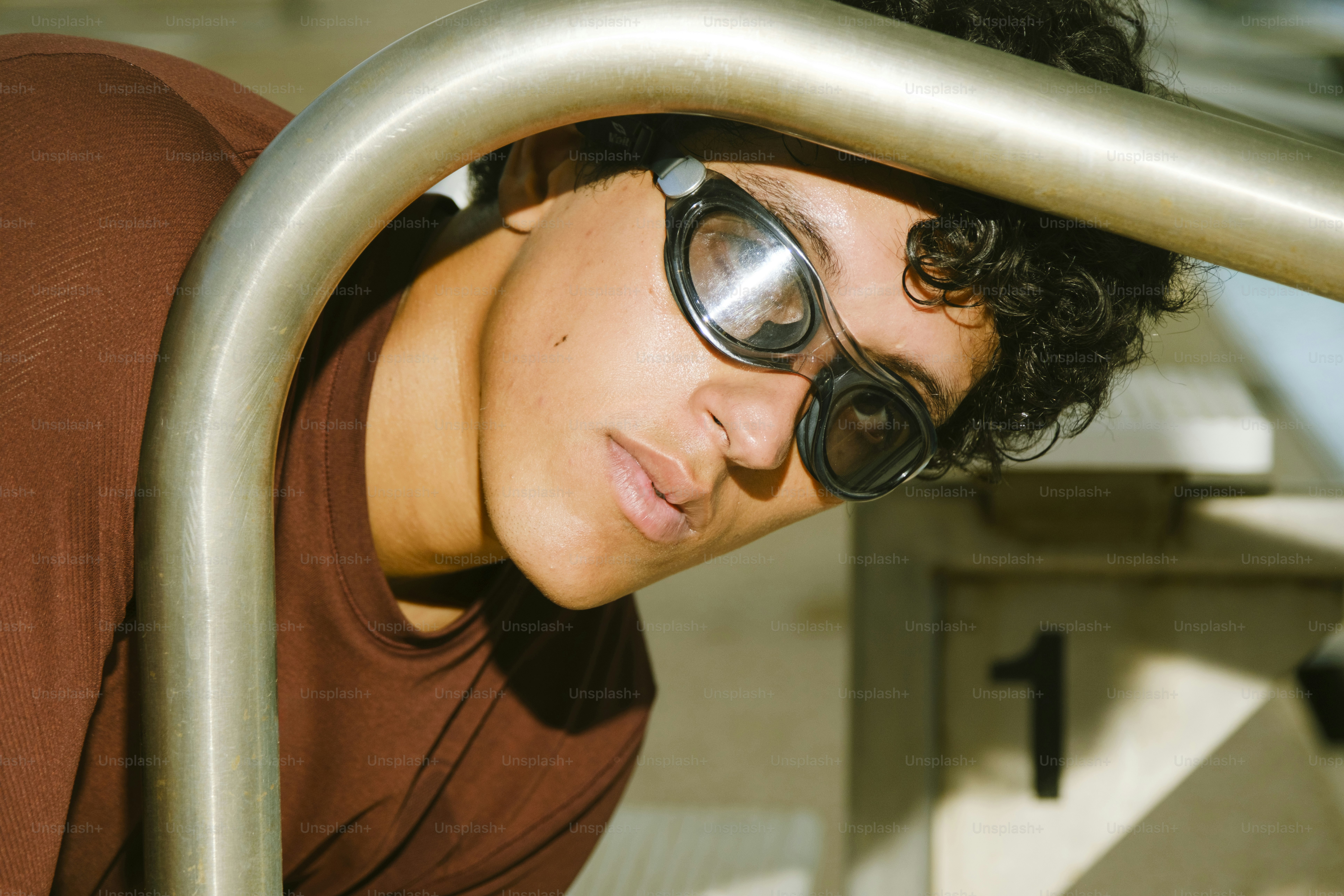 A person wearing swimming goggles and a brown shirt near a pool ladder