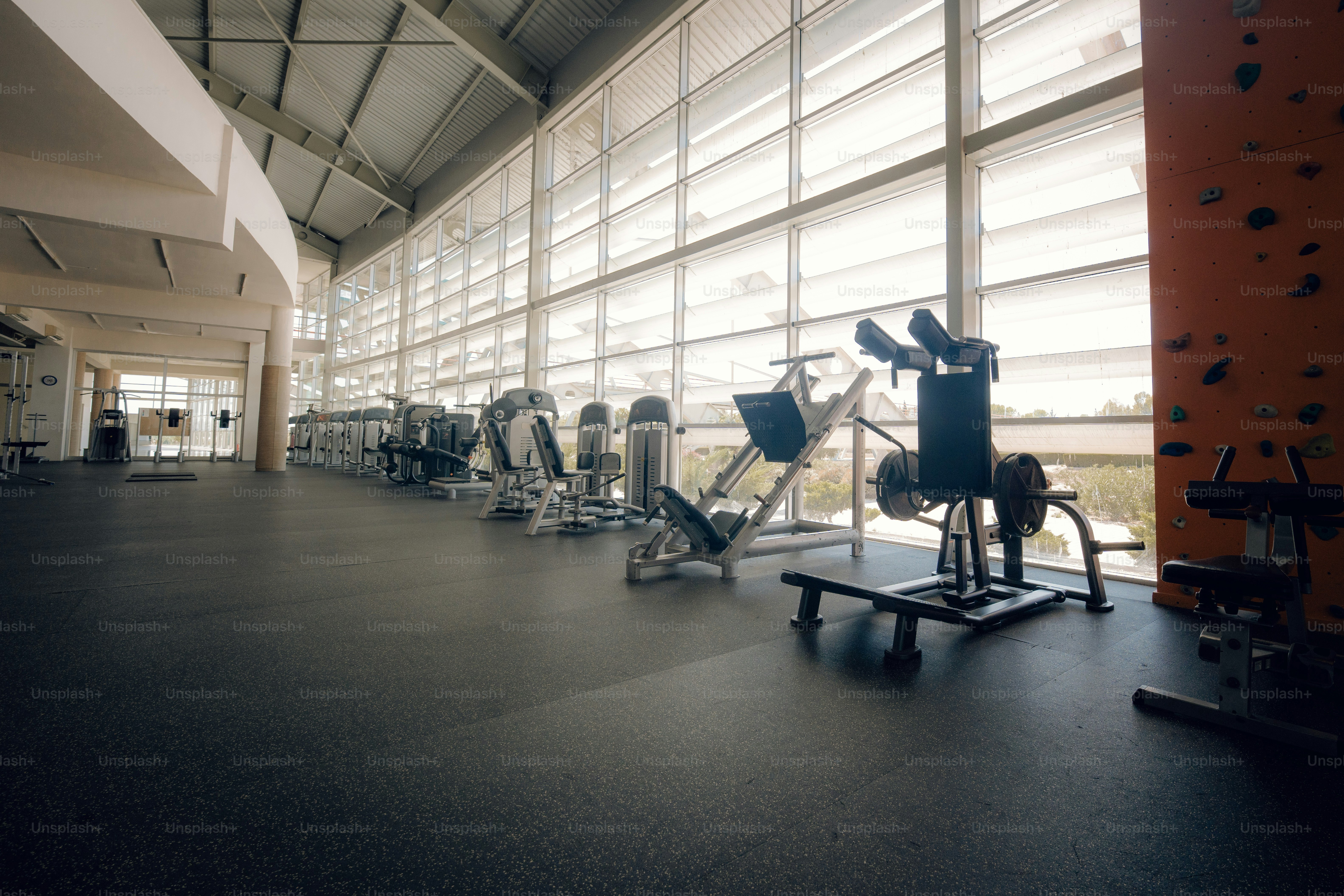 Exercise machines in a sunlit gym with a climbing wall and large windows