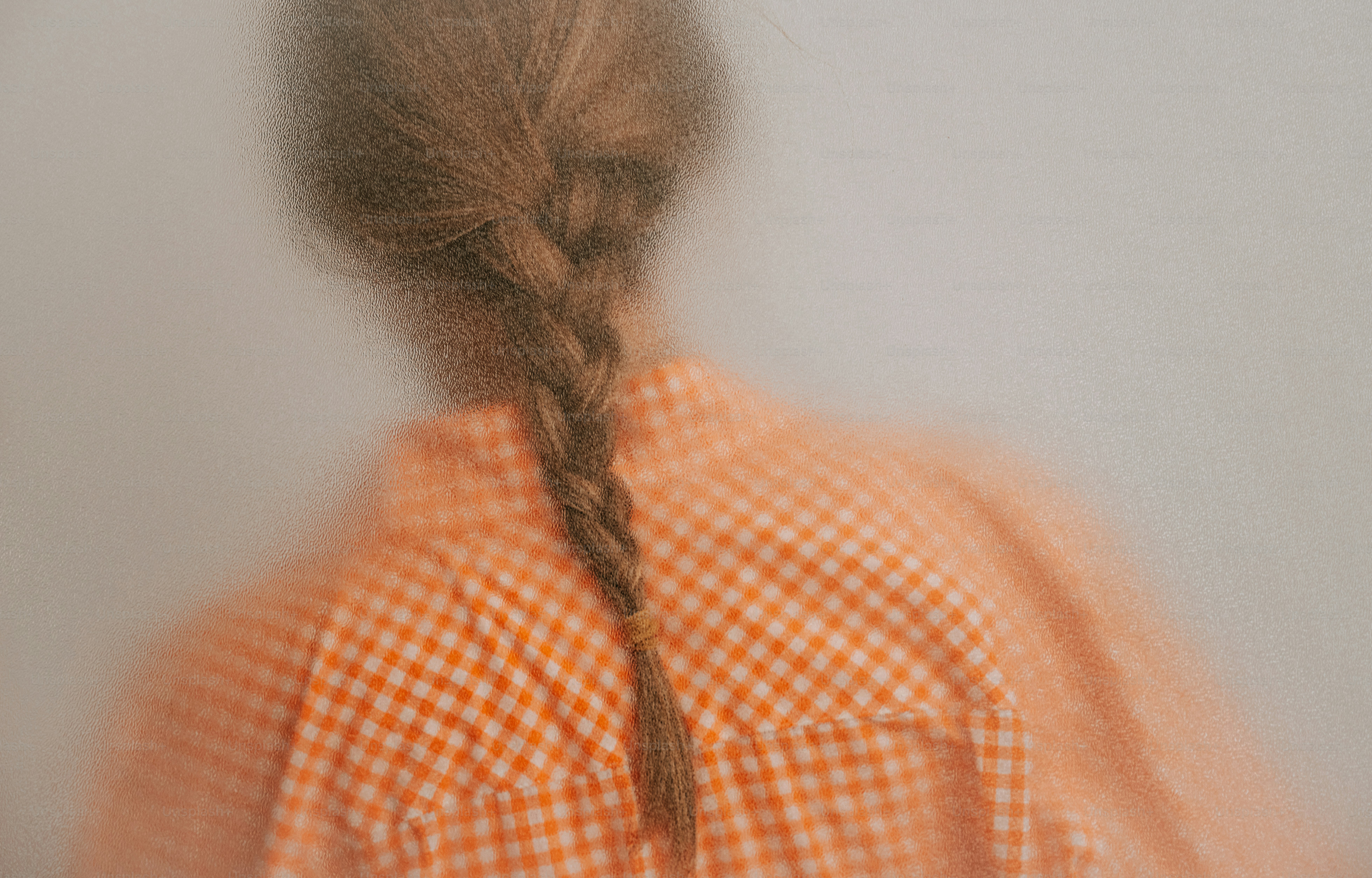 A person with a braided hairstyle wearing an orange gingham shirt behind frosted glass