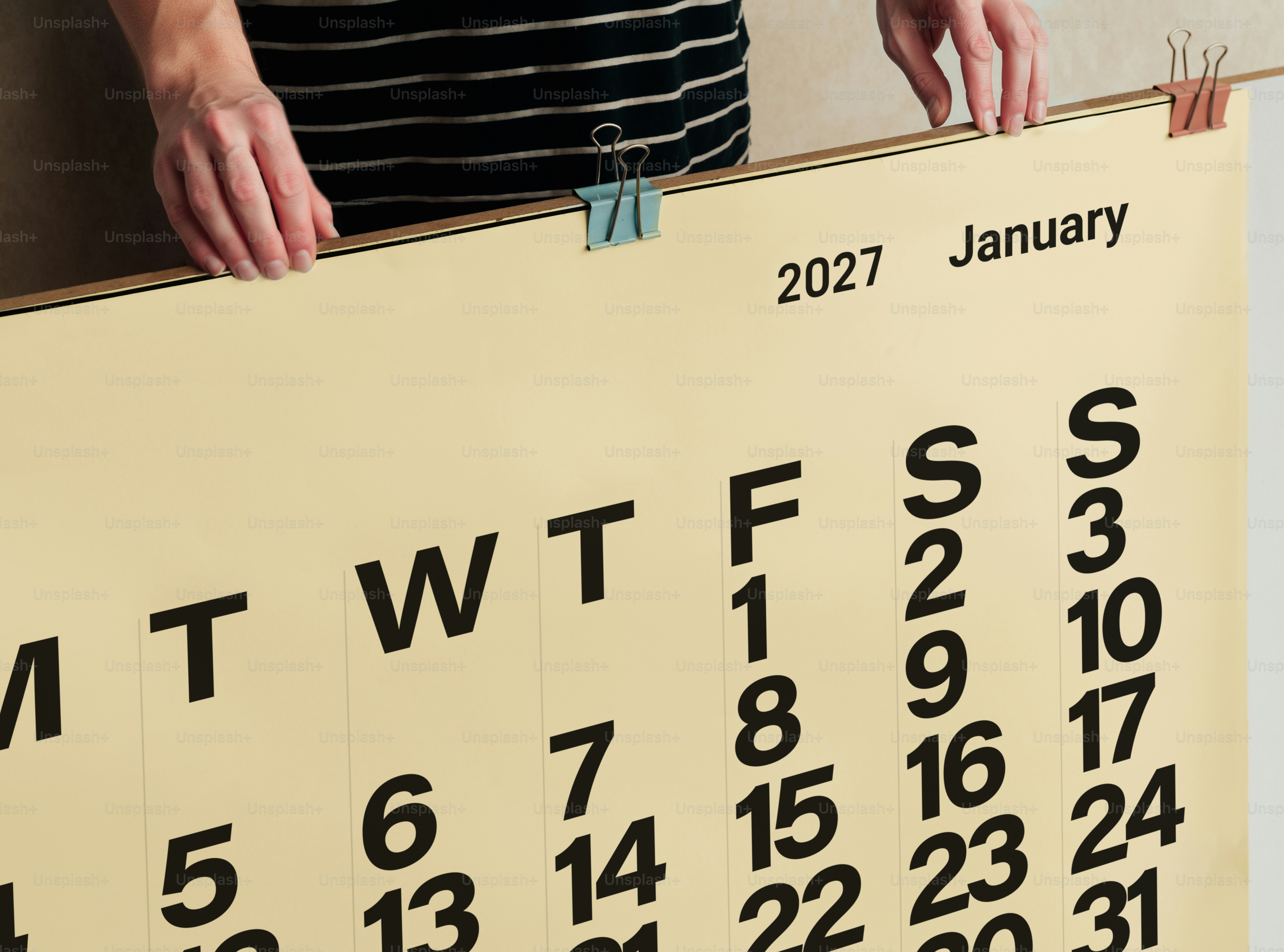 A person holding a large wall calendar for January 2027 with metal clips
