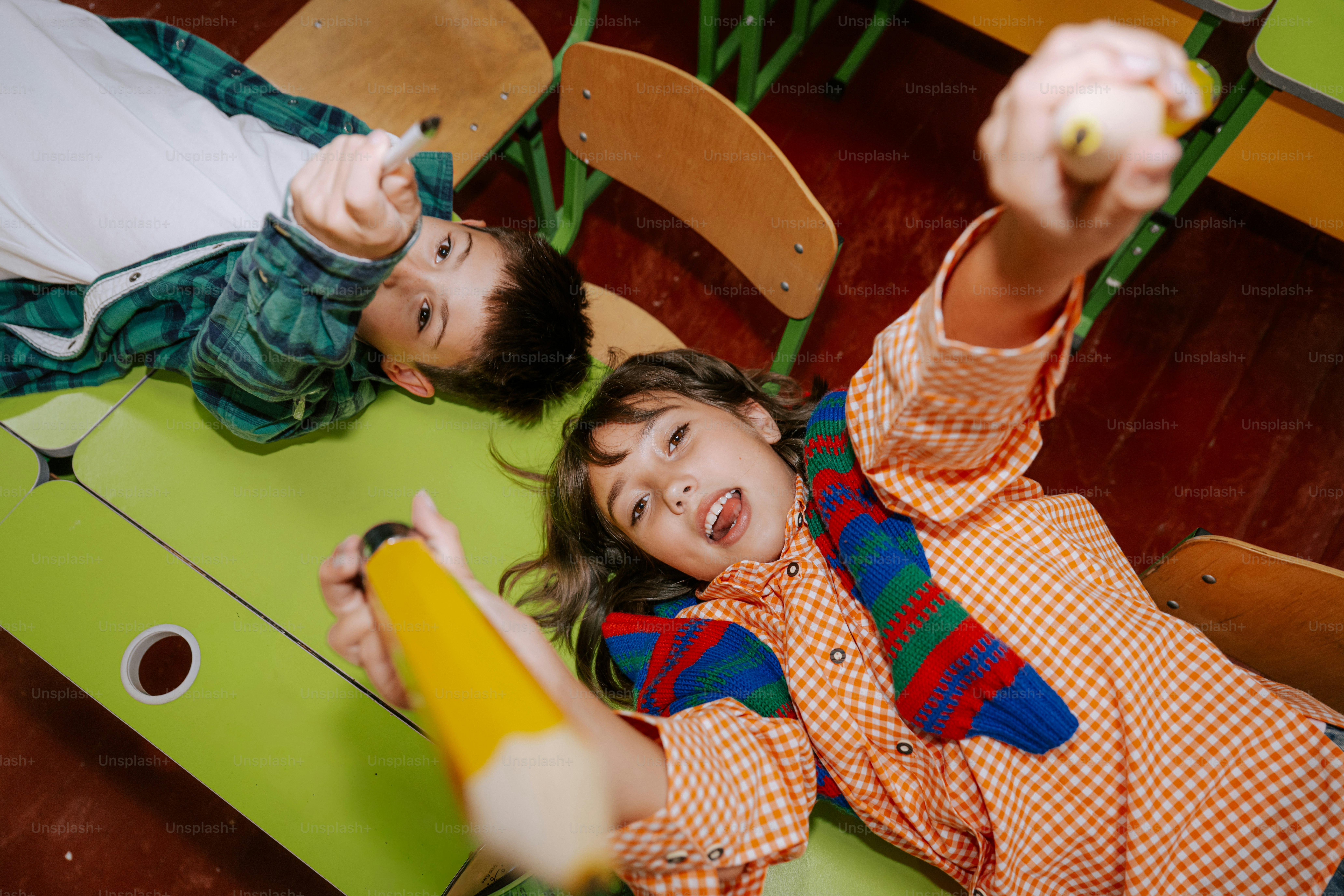 Two children lying on green desks holding pencils toward the camera