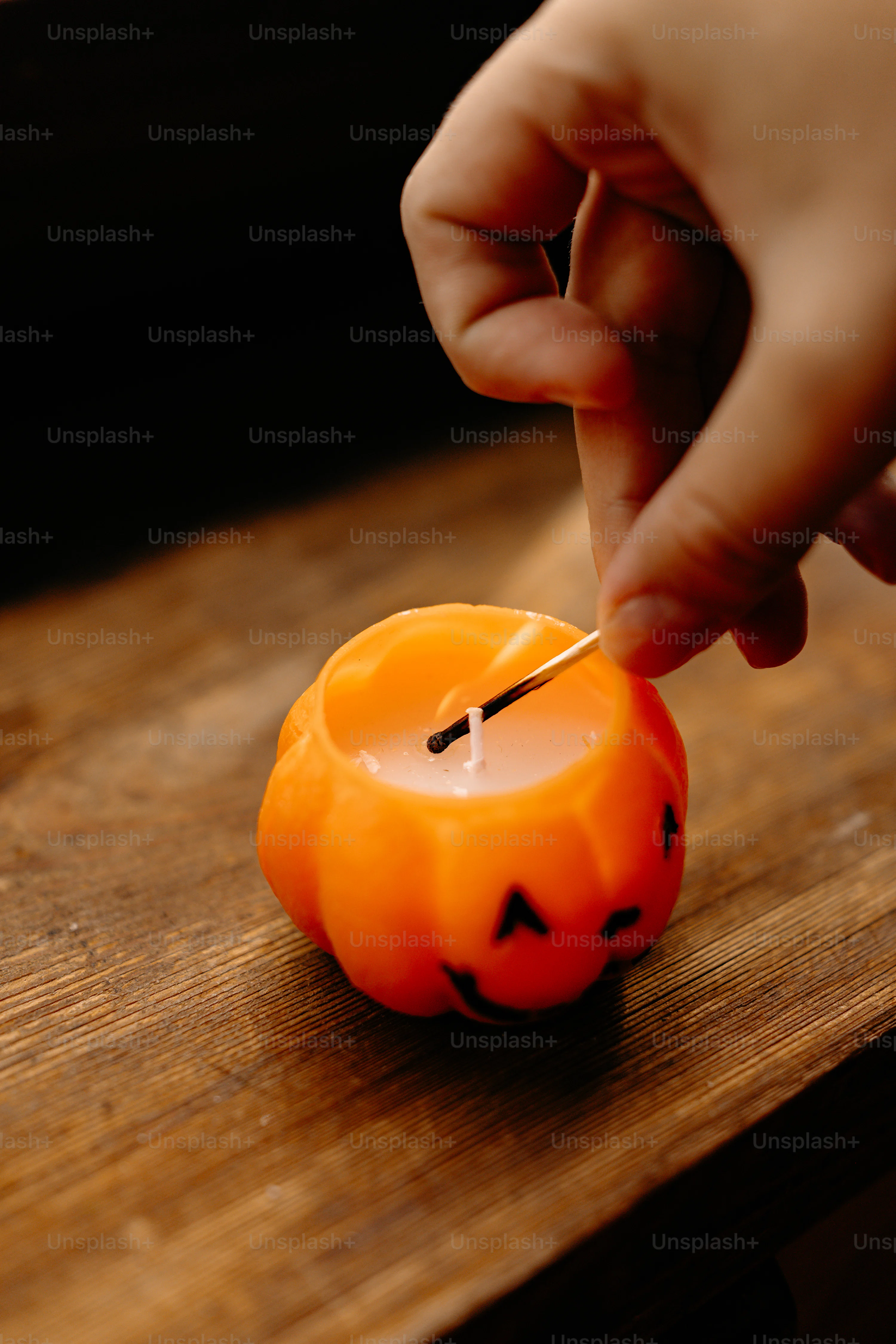 A hand lighting a pumpkin-shaped candle with a match on a wooden surface