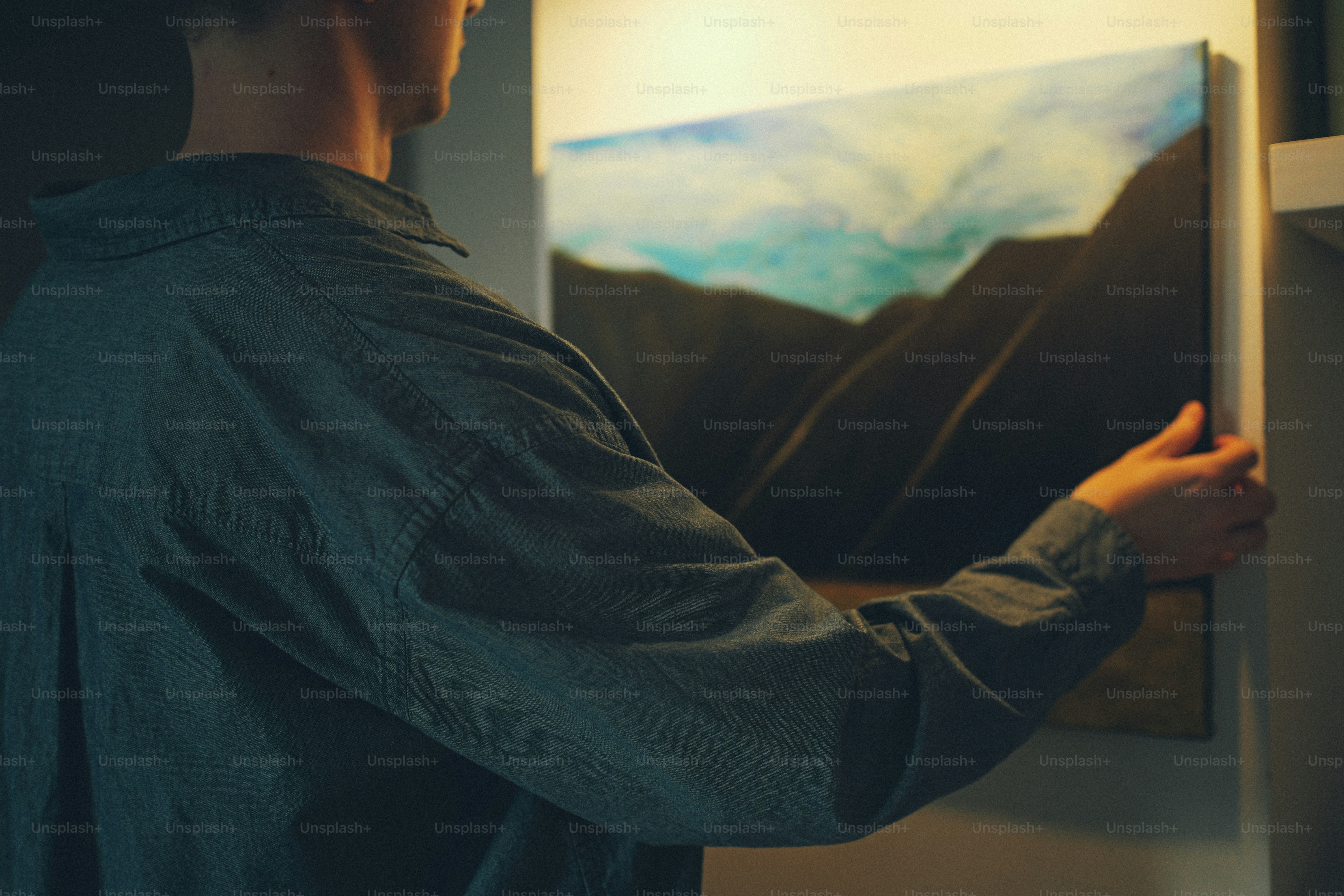 A person in a grey shirt hanging a landscape painting on a wall