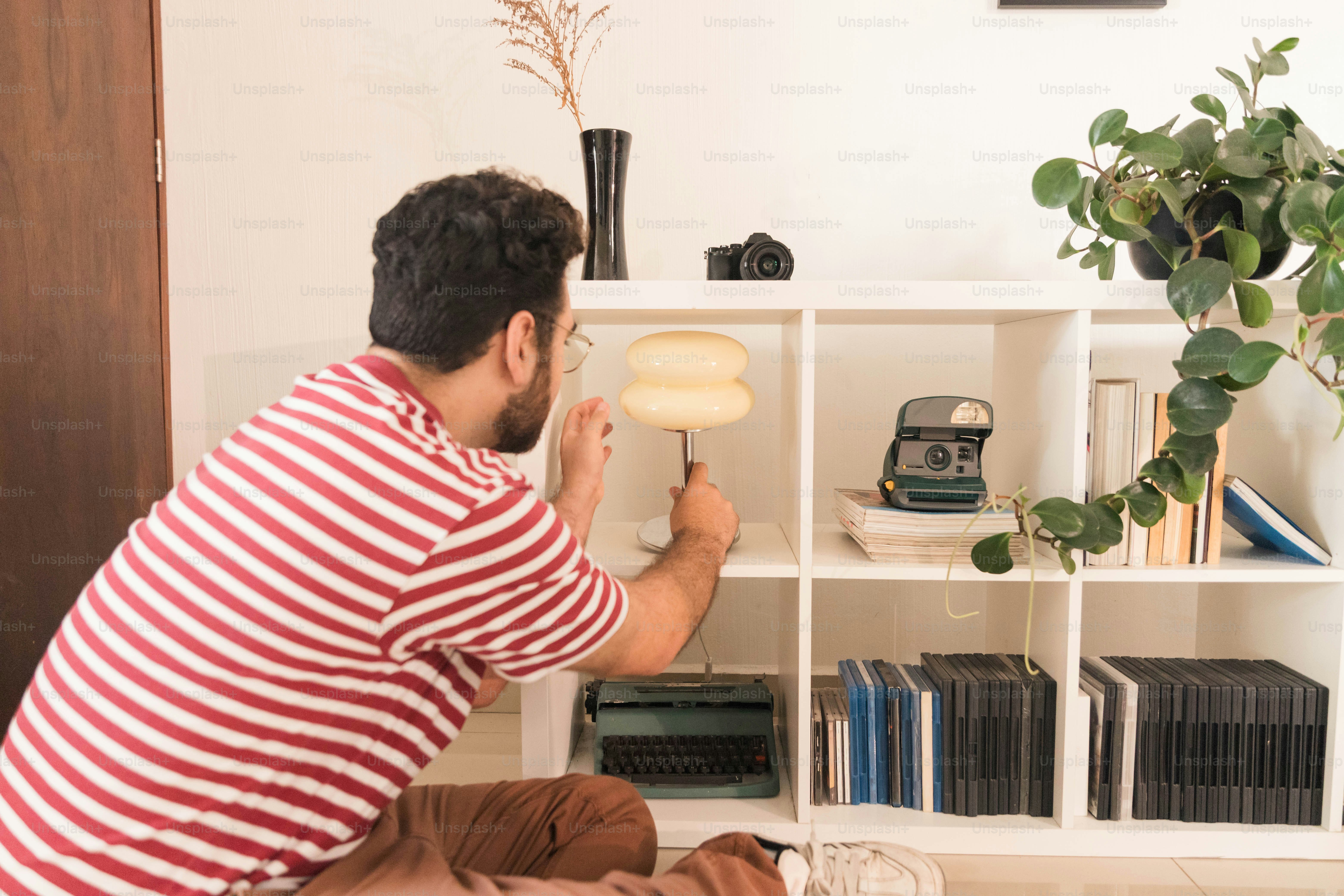 A man in a striped shirt adjusting a lamp on a white bookshelf