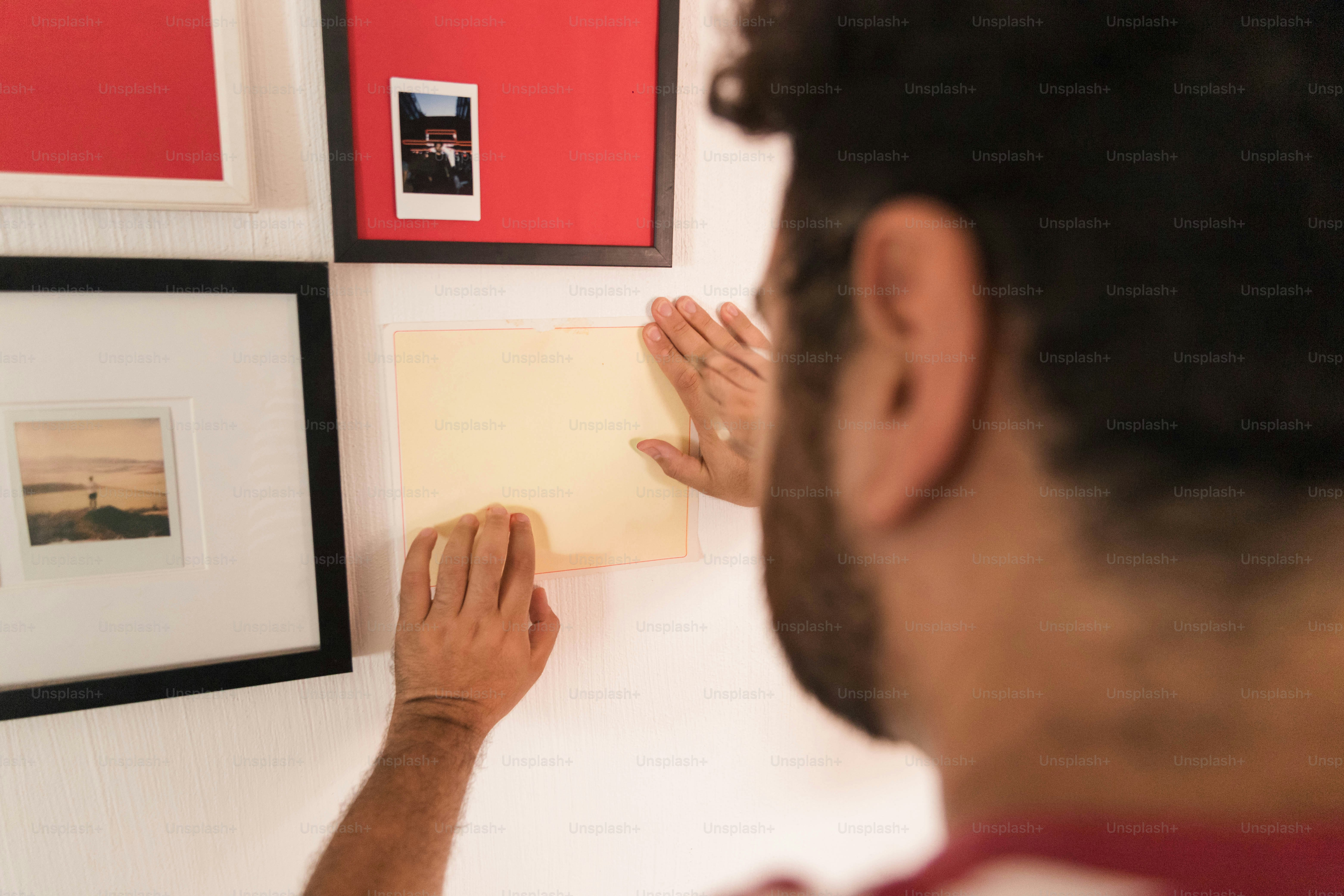 A person attaching a blank rectangular paper to a white wall with framed photos