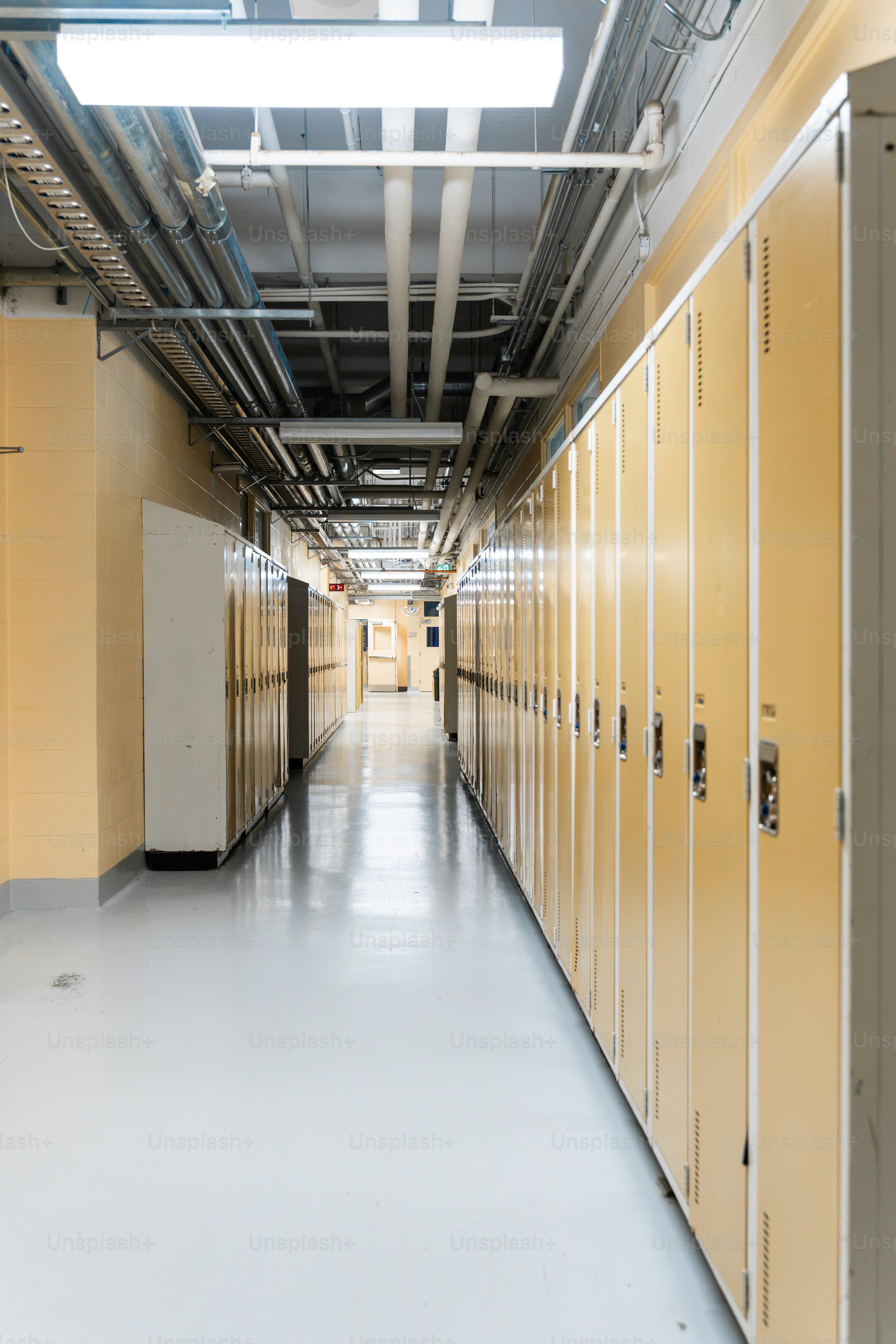 Un long couloir d’école bordé de casiers en métal jaune et de tuyaux au plafond apparents