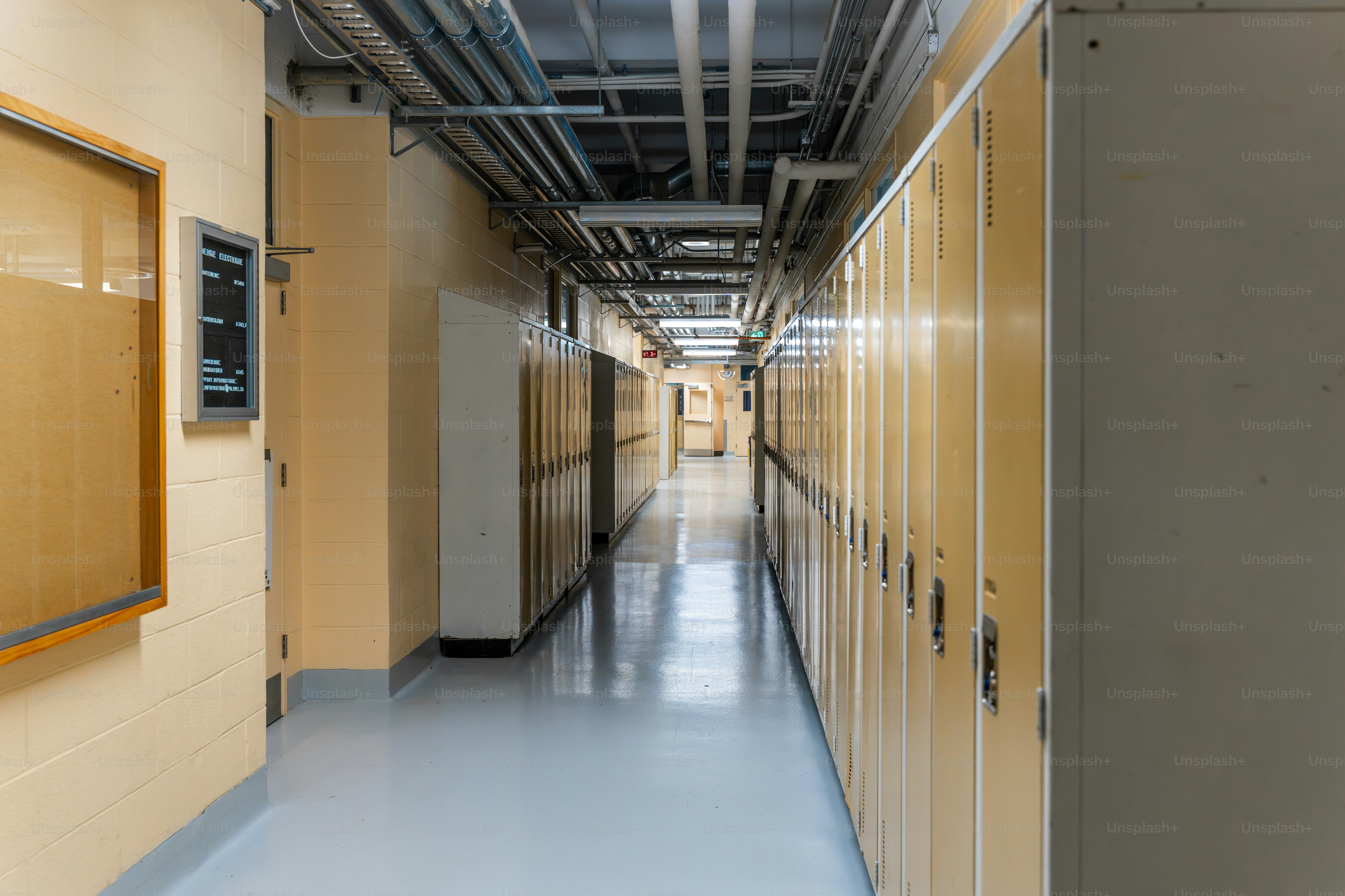 Un long couloir d’école bordé de rangées de casiers métalliques jaunes et de tuyaux exposés