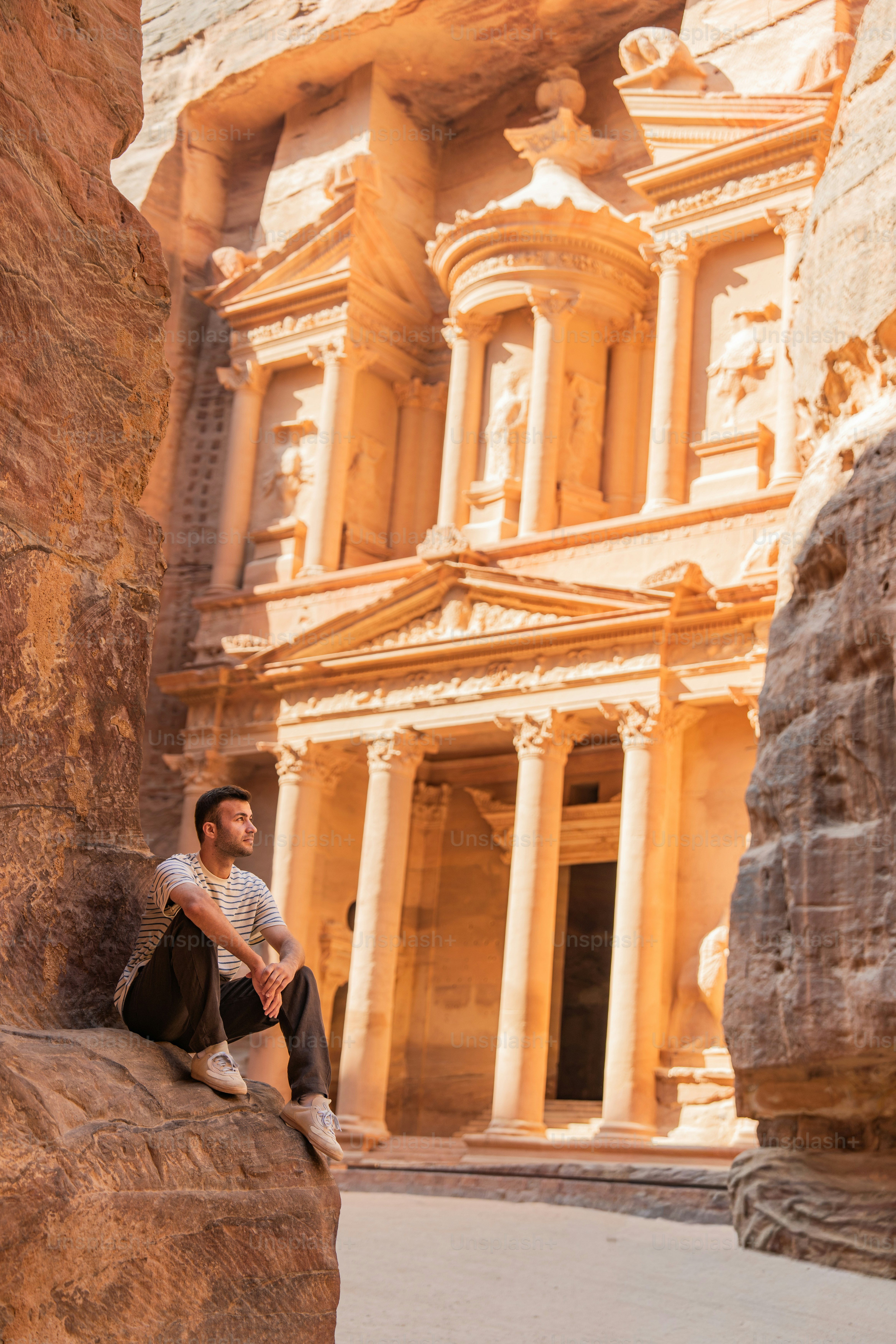 A man in a striped shirt sitting on a rock before the Treasury in Petra