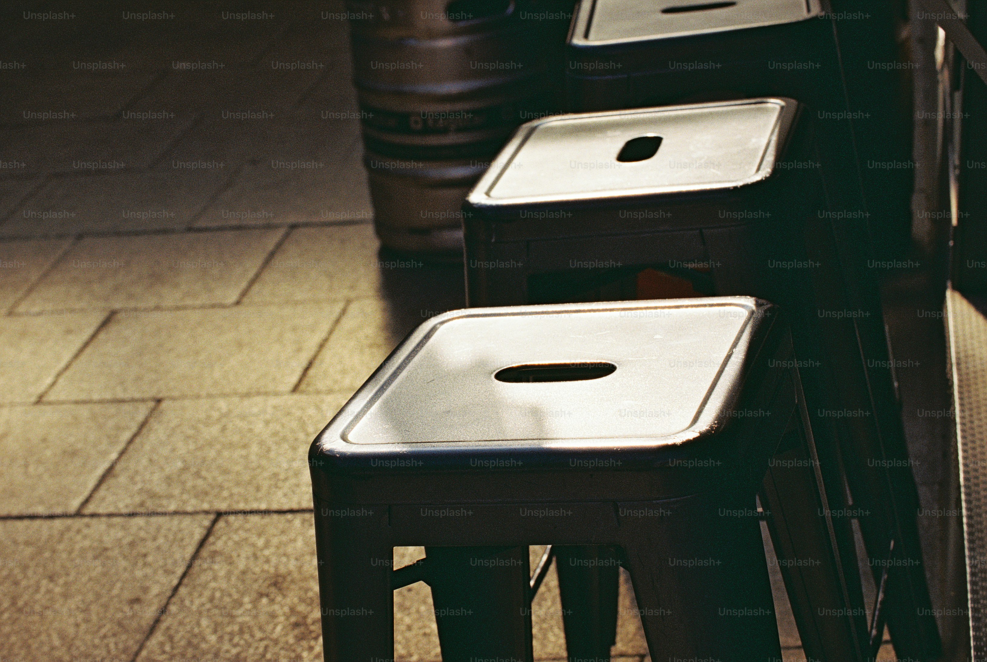 Dark metal stools and a steel keg on paved ground in warm light