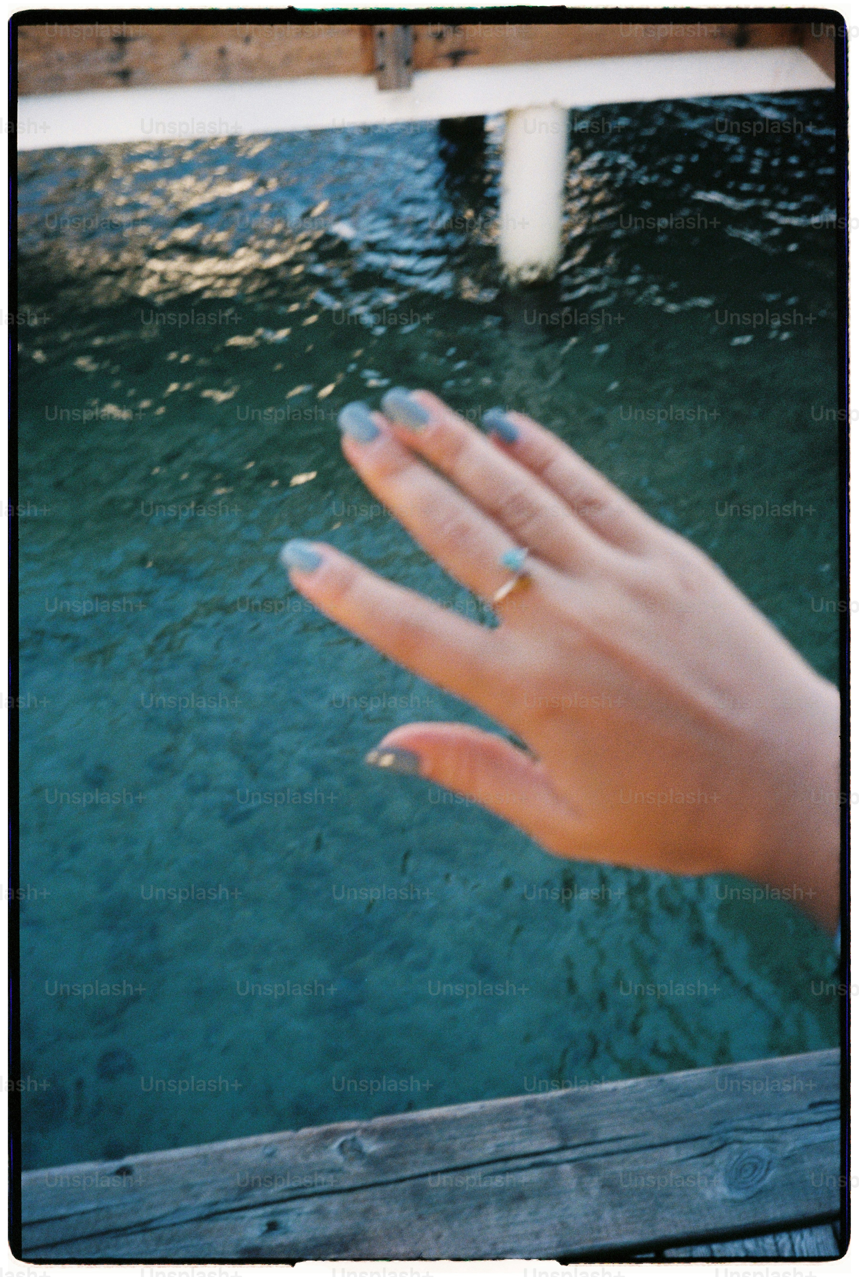 A hand with blue nail polish and a ring over turquoise water
