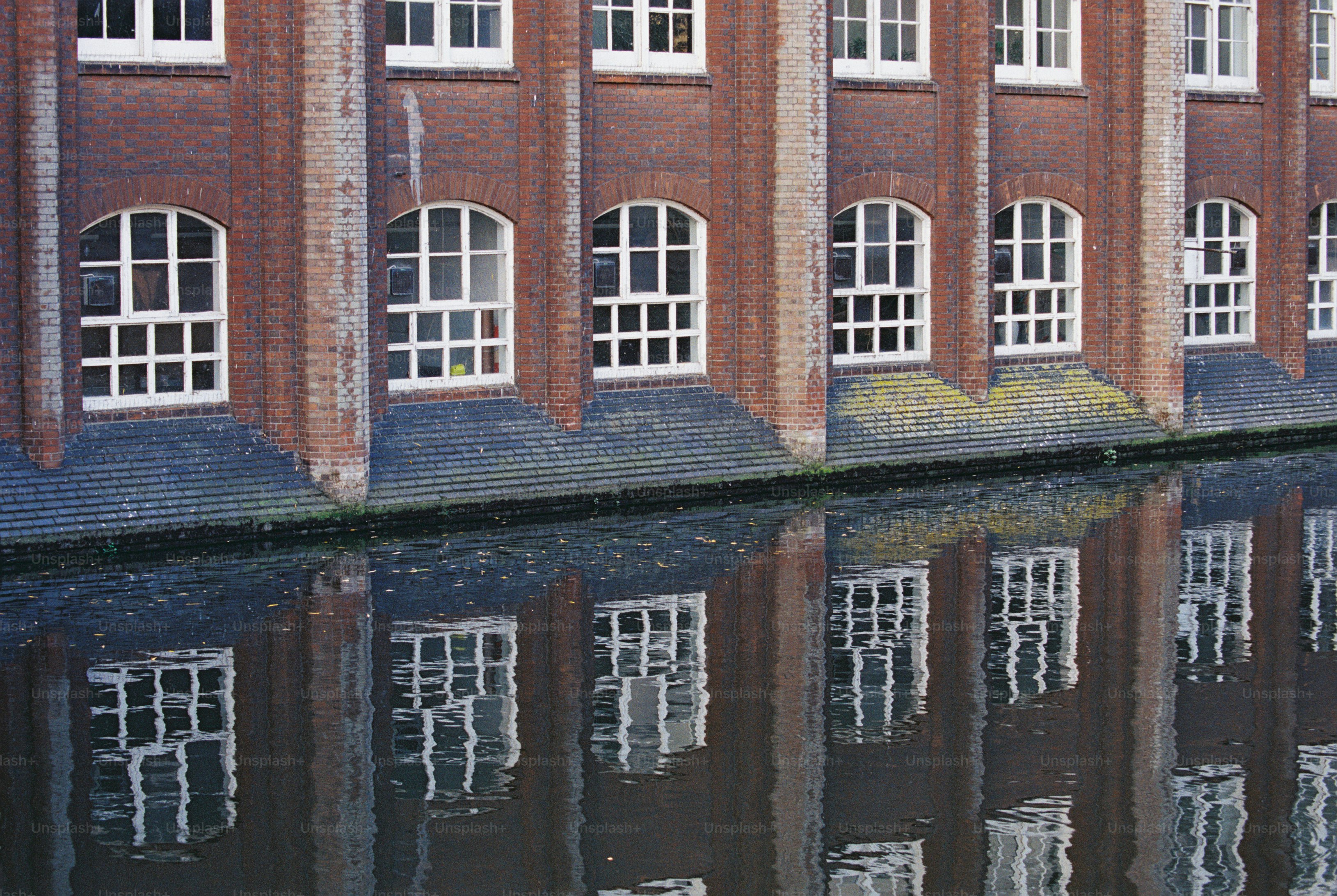 A red brick building with arched white-framed windows reflecting in dark canal water