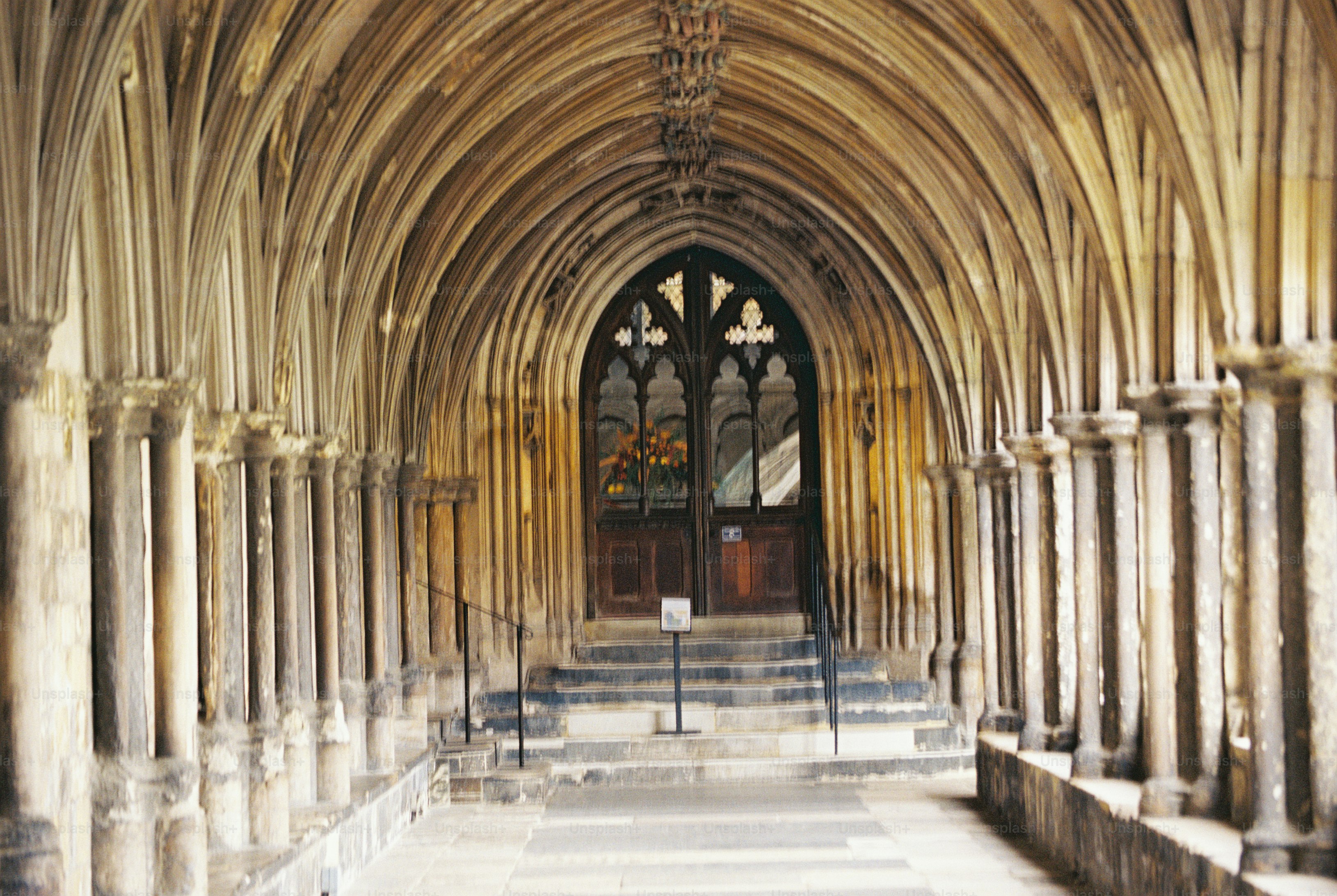 A vaulted stone cloister walkway with slender columns leading to a carved wooden door