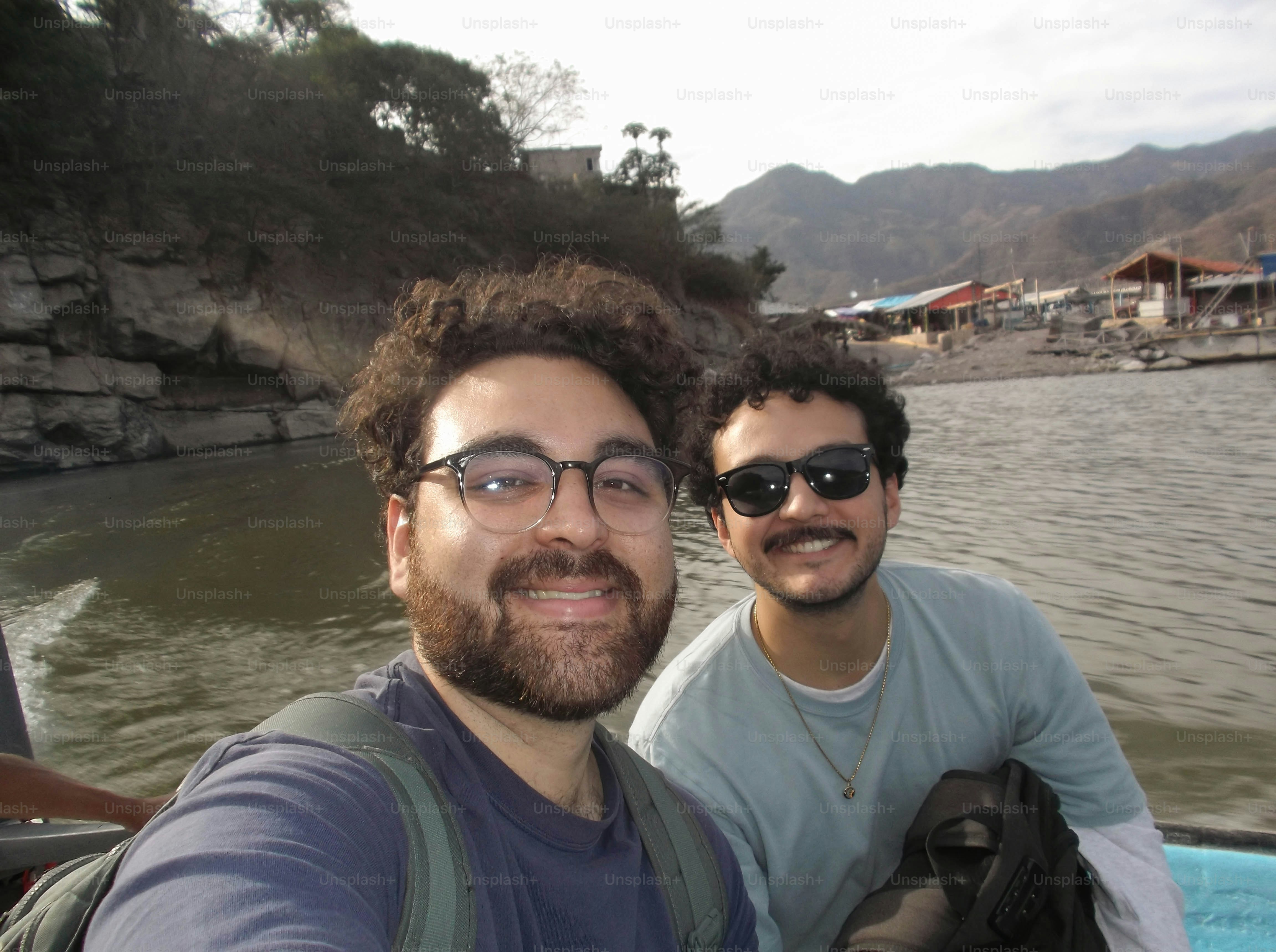 Two men with beards smiling on a boat on a river near mountains