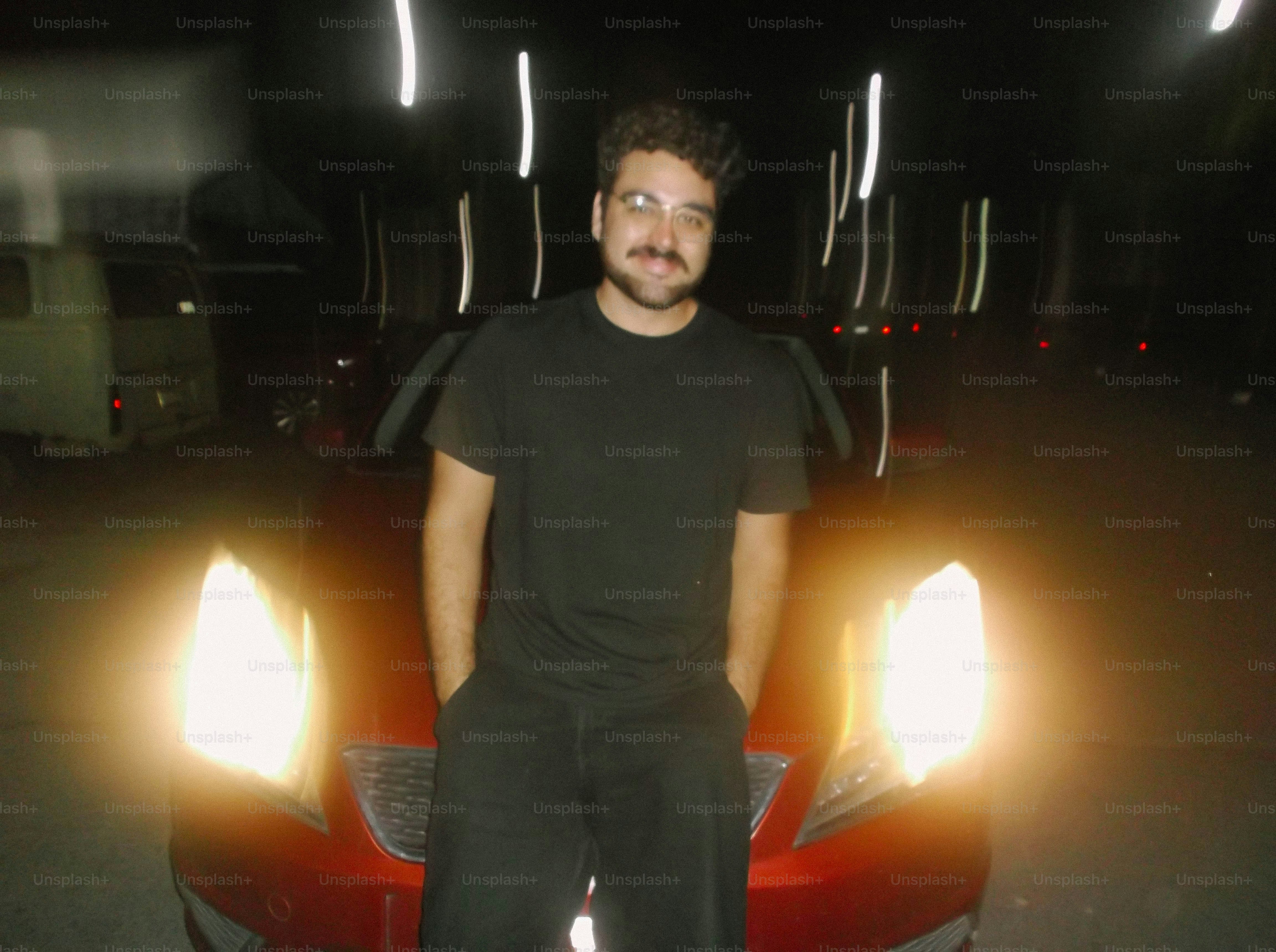 A man in a black t-shirt leaning against a red car at night