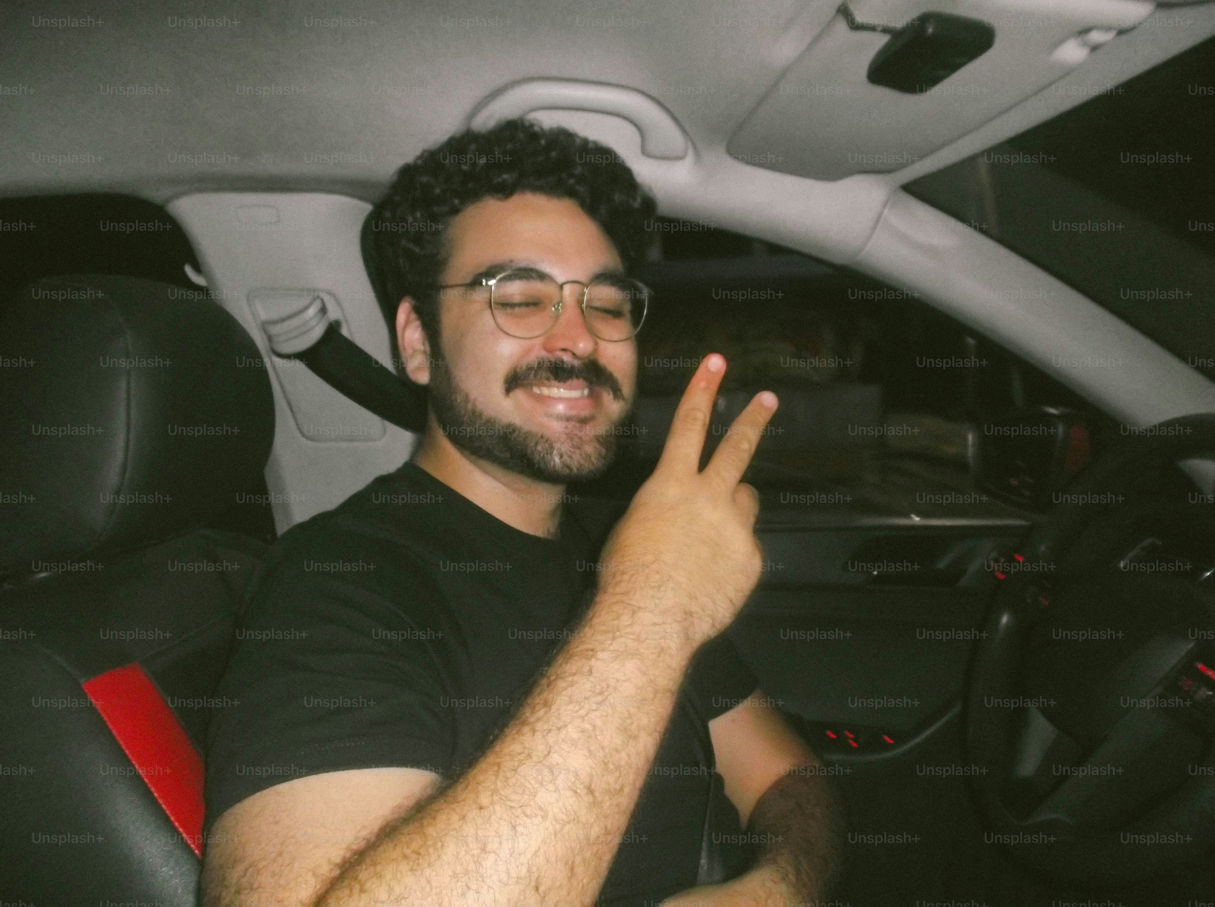 A man with a mustache and glasses making a peace sign in a car