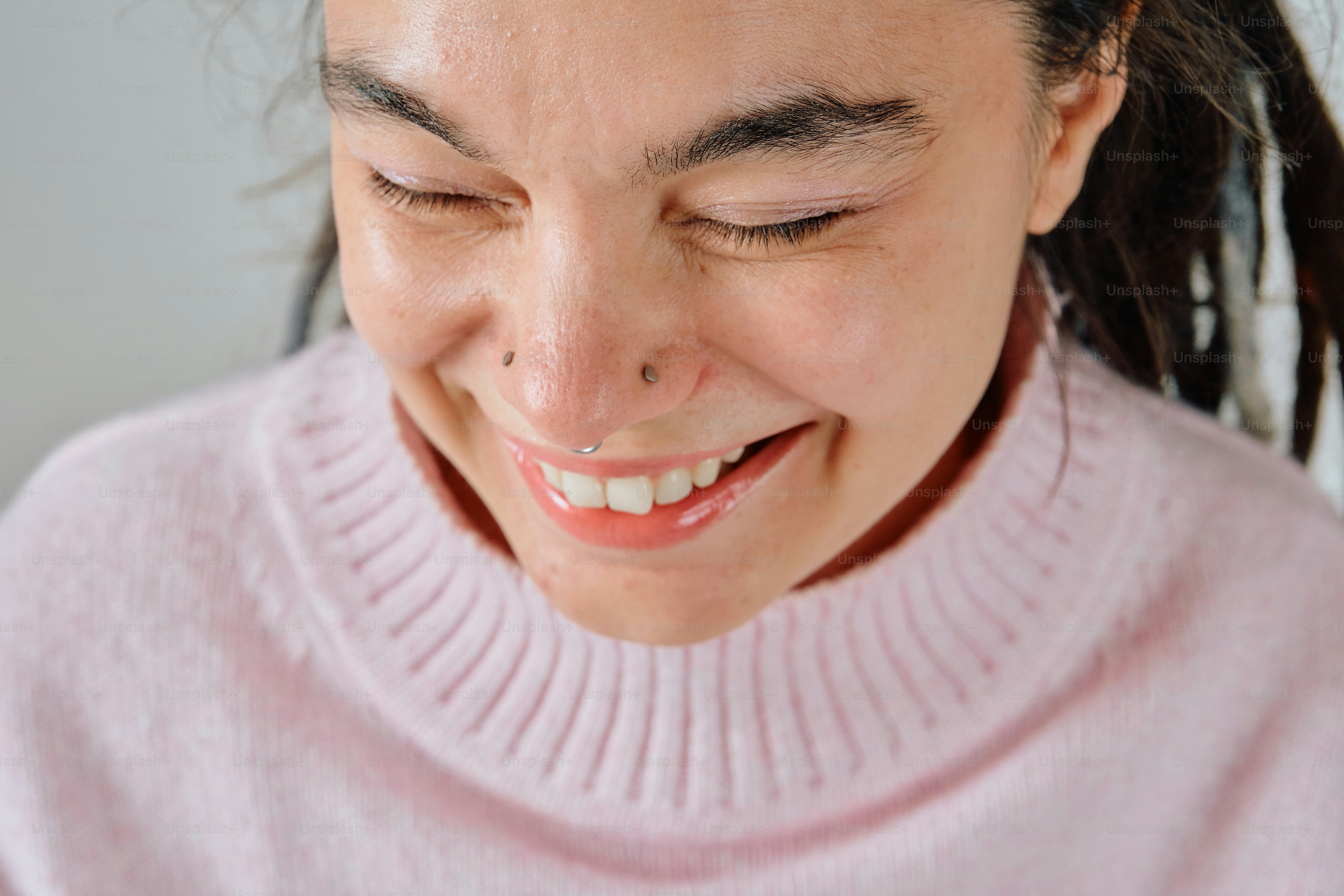A person with dreadlocks and a nose piercing laughing in a pink sweater