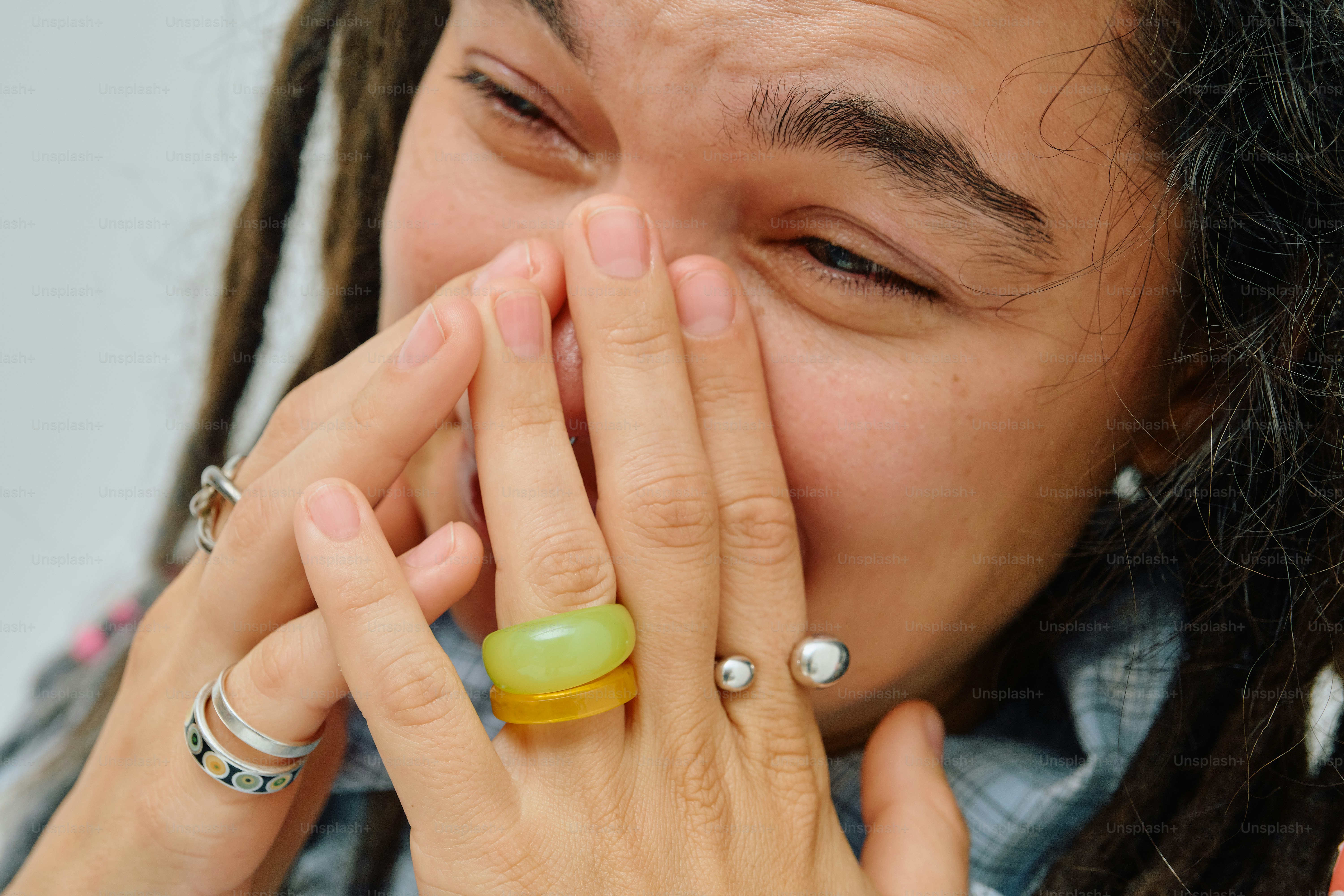 A person with dreadlocks covering their mouth with hands adorned with colorful rings