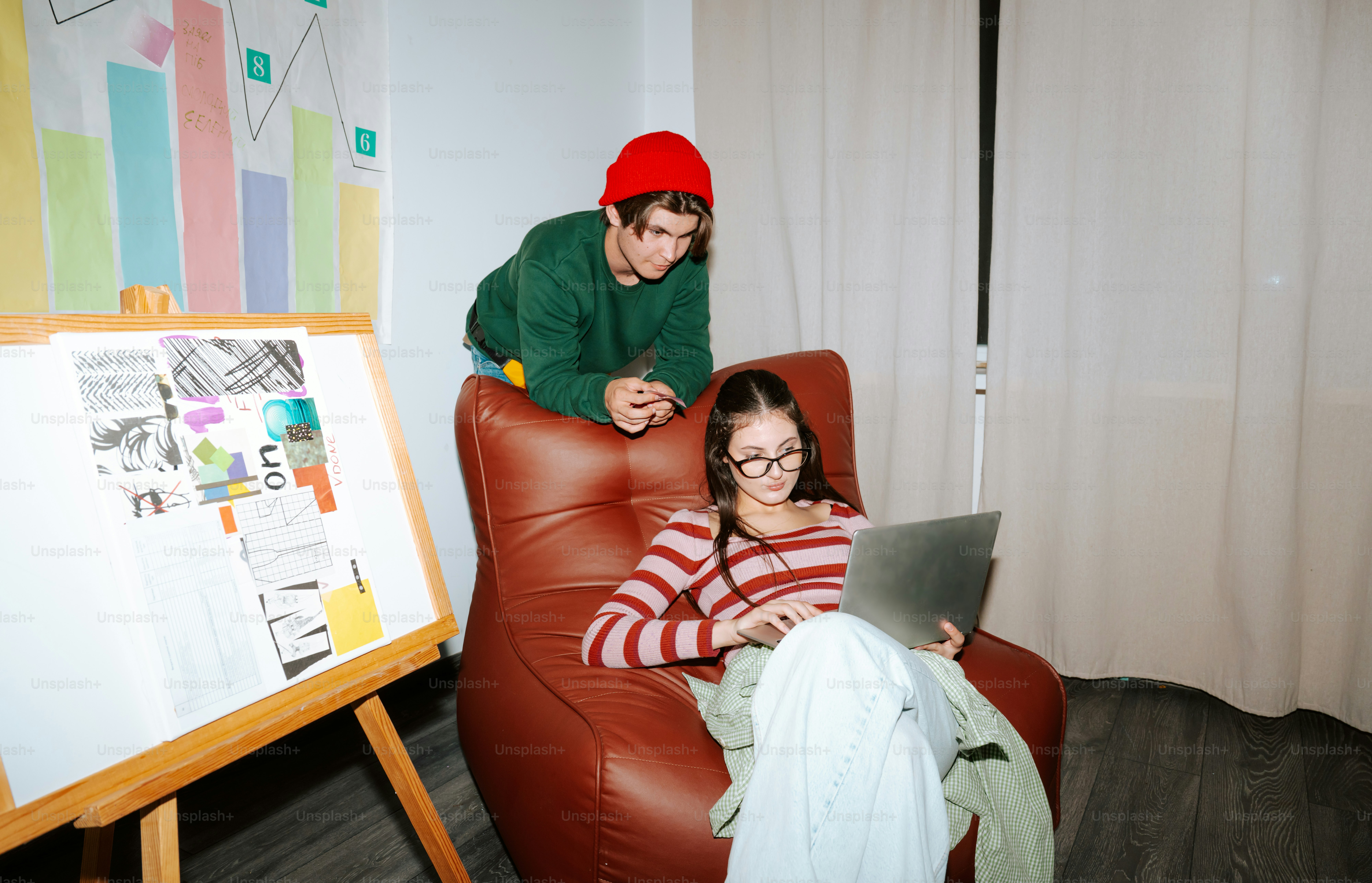 A woman using a laptop in an armchair beside a man in a red beanie