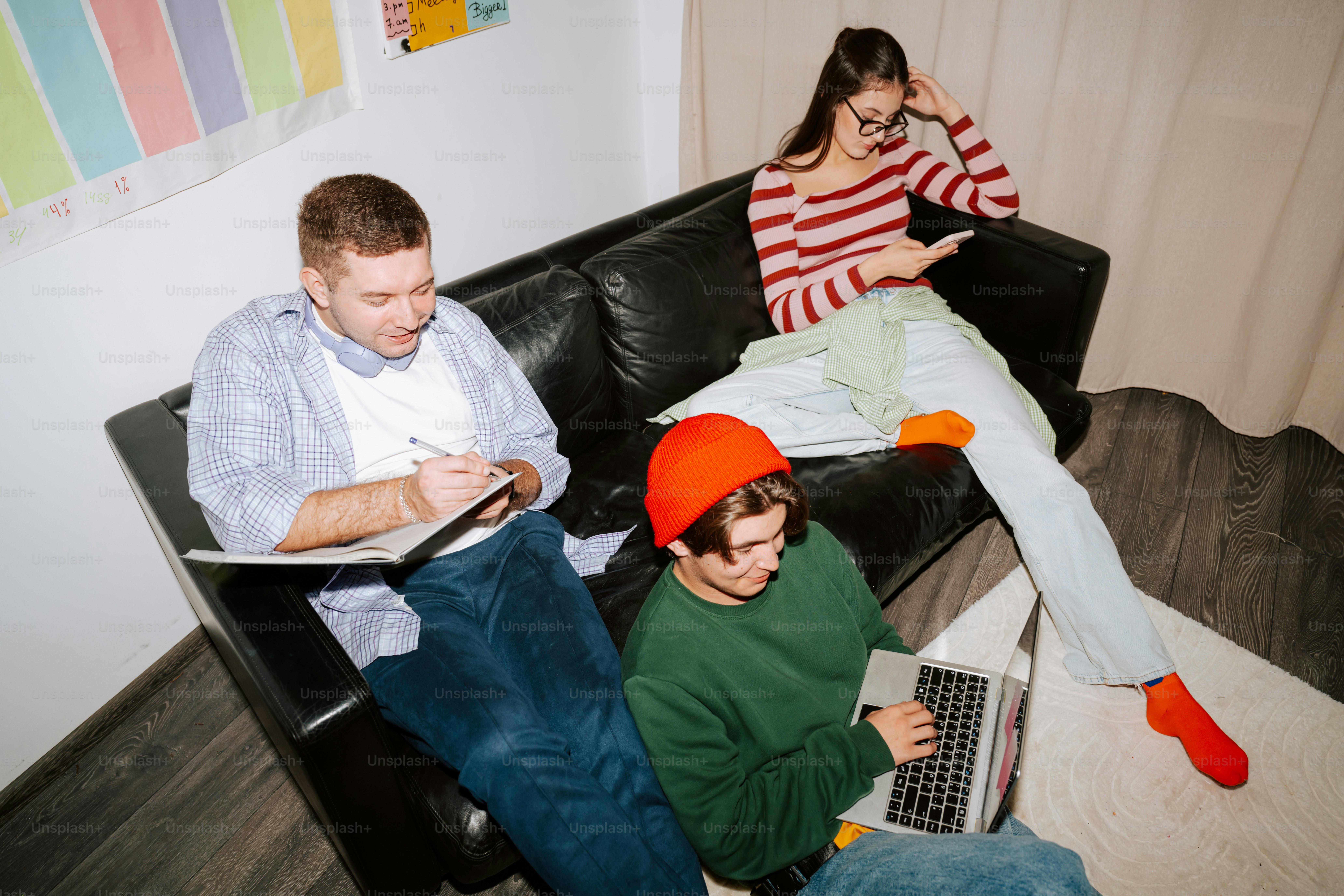Three people on a black leather sofa, one writing, one using a laptop