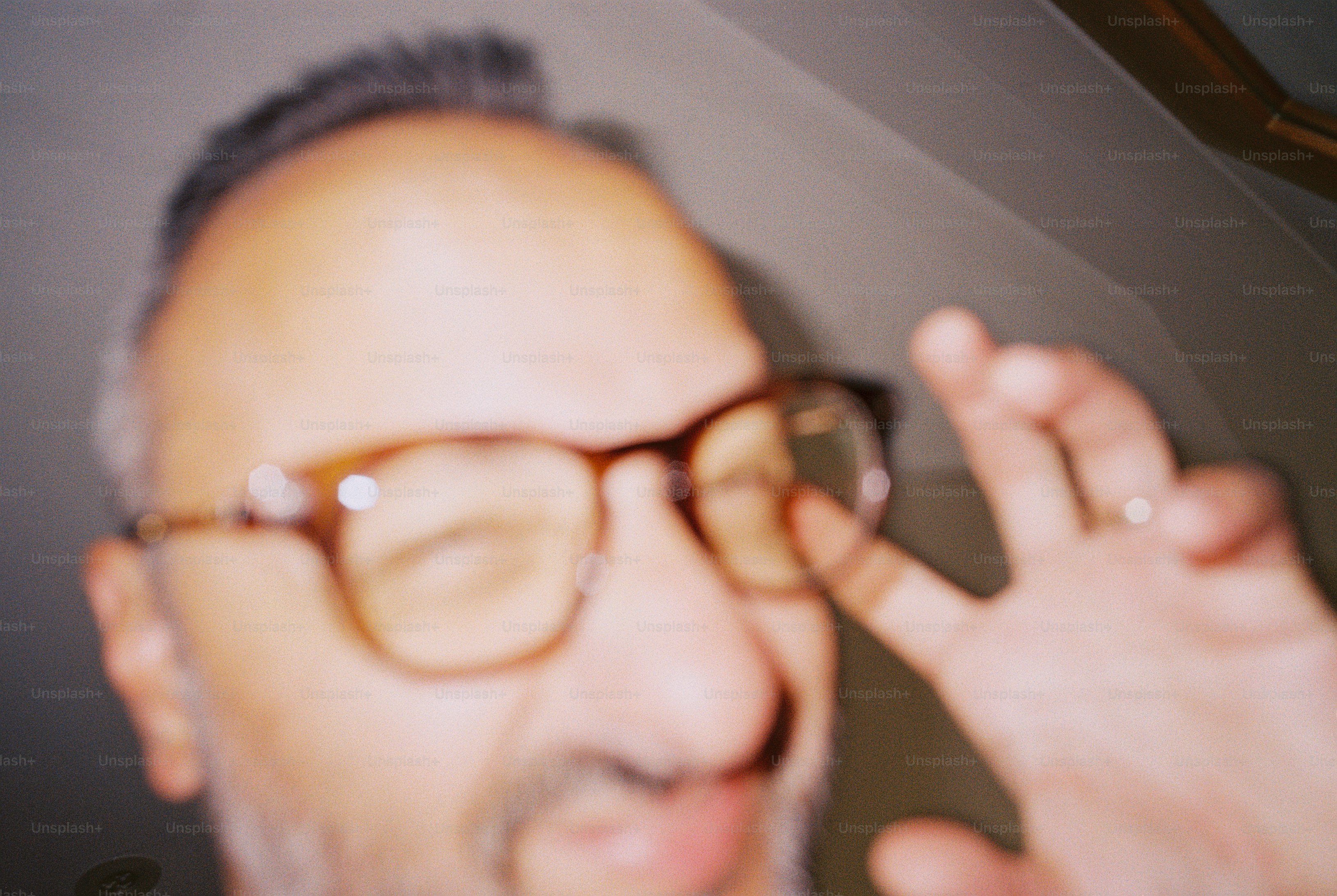 A blurred portrait of a man in tortoiseshell glasses with two fingers raised