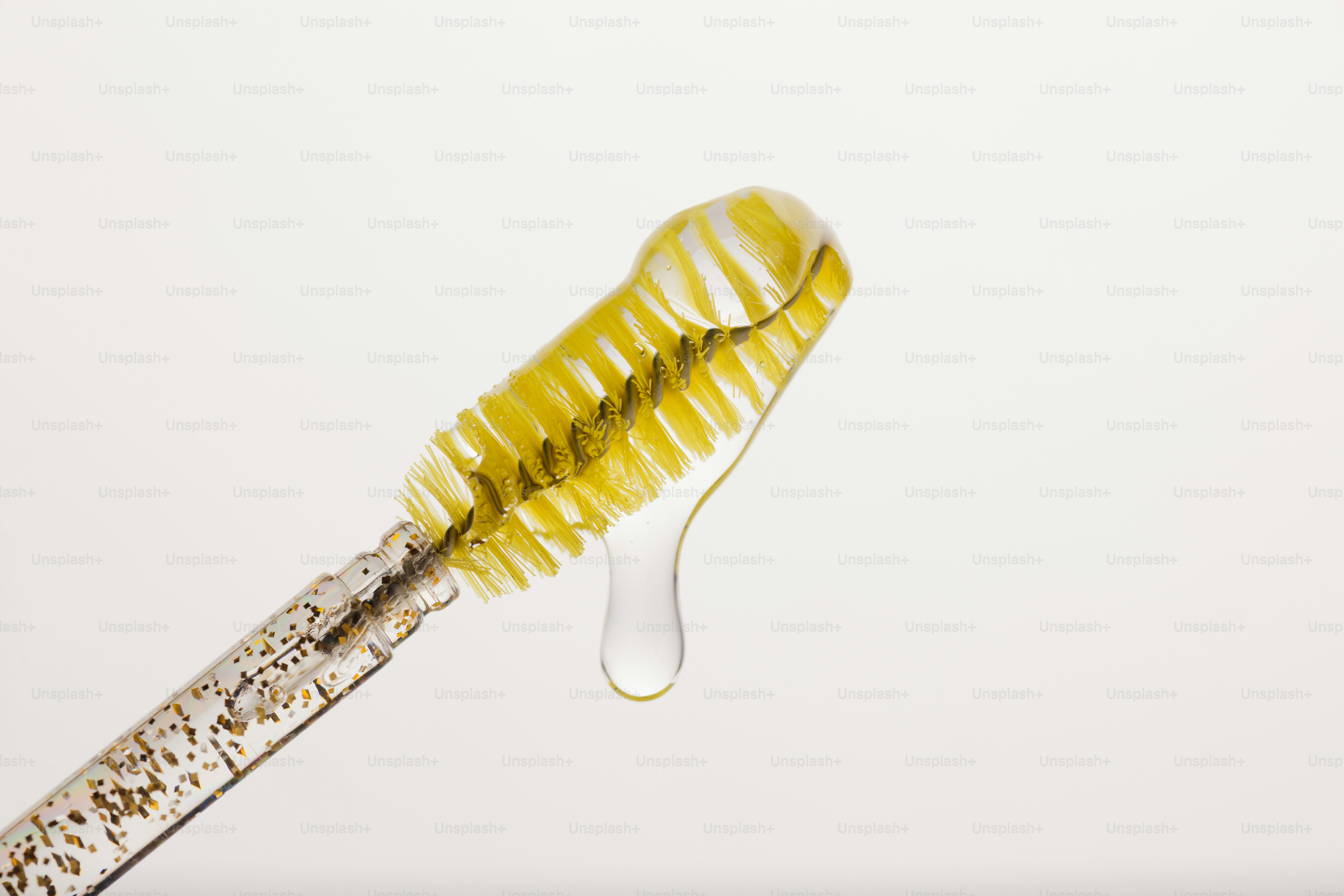 A yellow mascara wand with a clear drop of liquid dripping from bristles