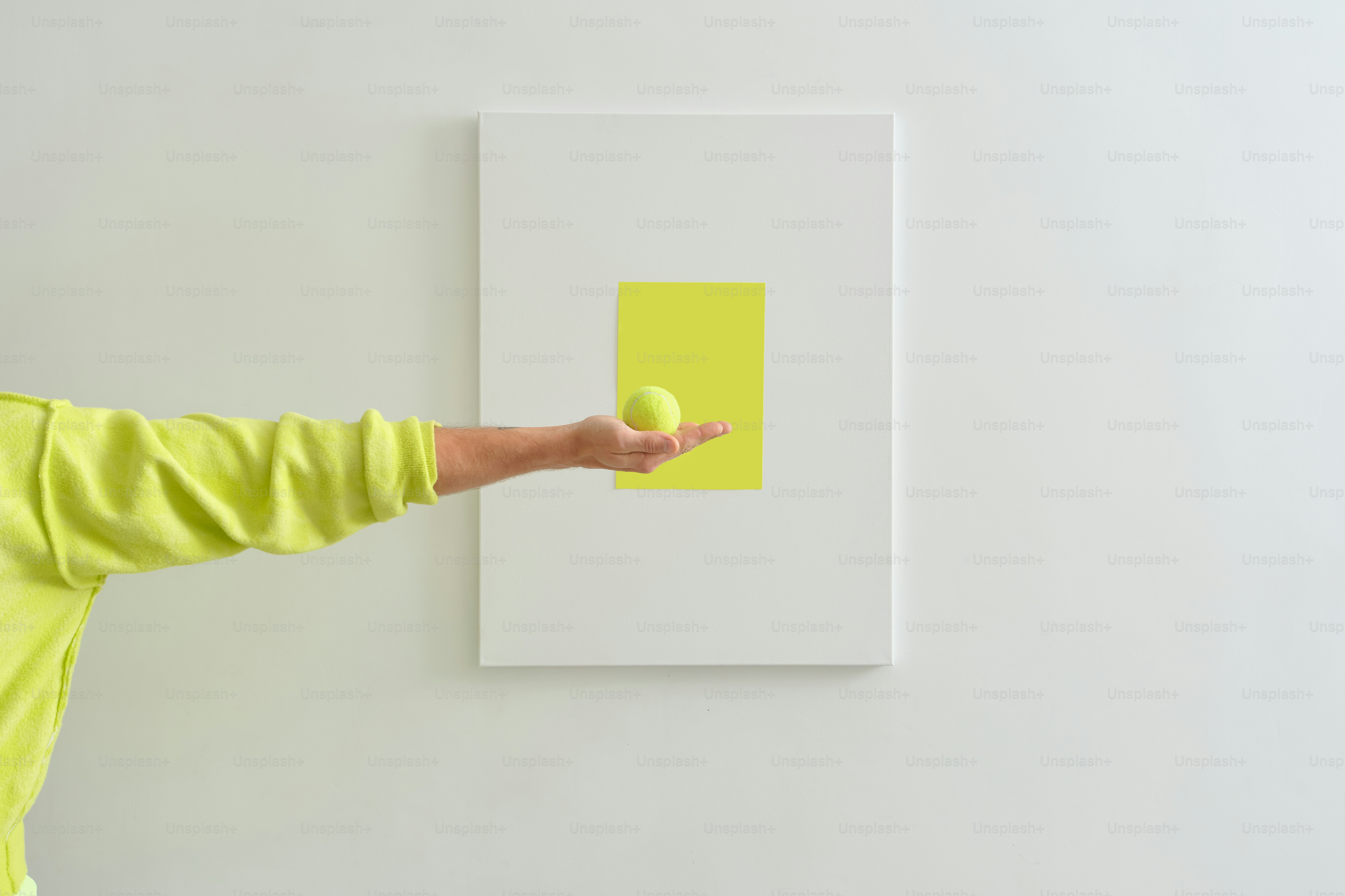 ネオンイエローのスウェットシャツを着て、白い壁にテニスボールを持った人物