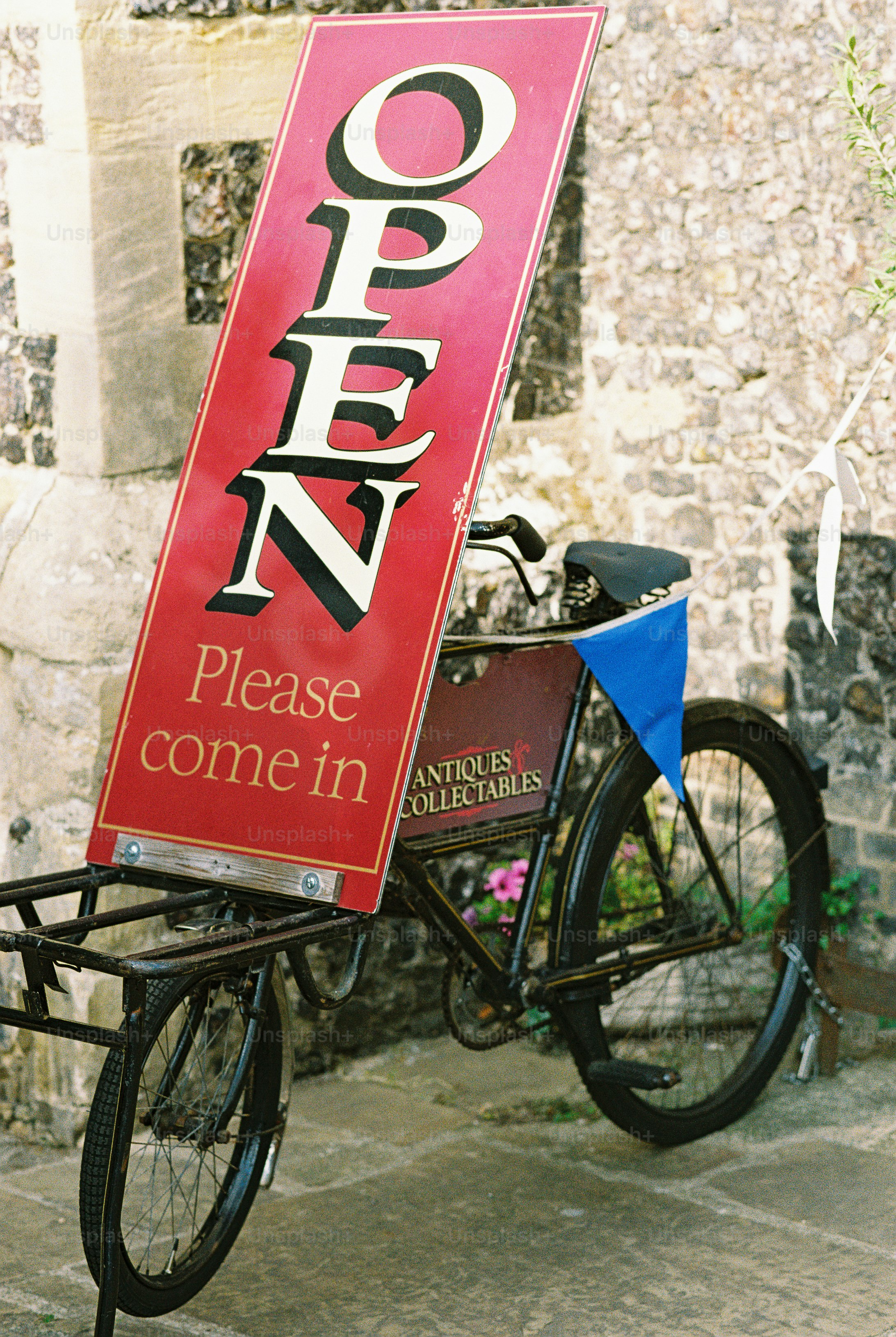A vintage bicycle with a red sign reading OPEN Please come in parked against a stone wall