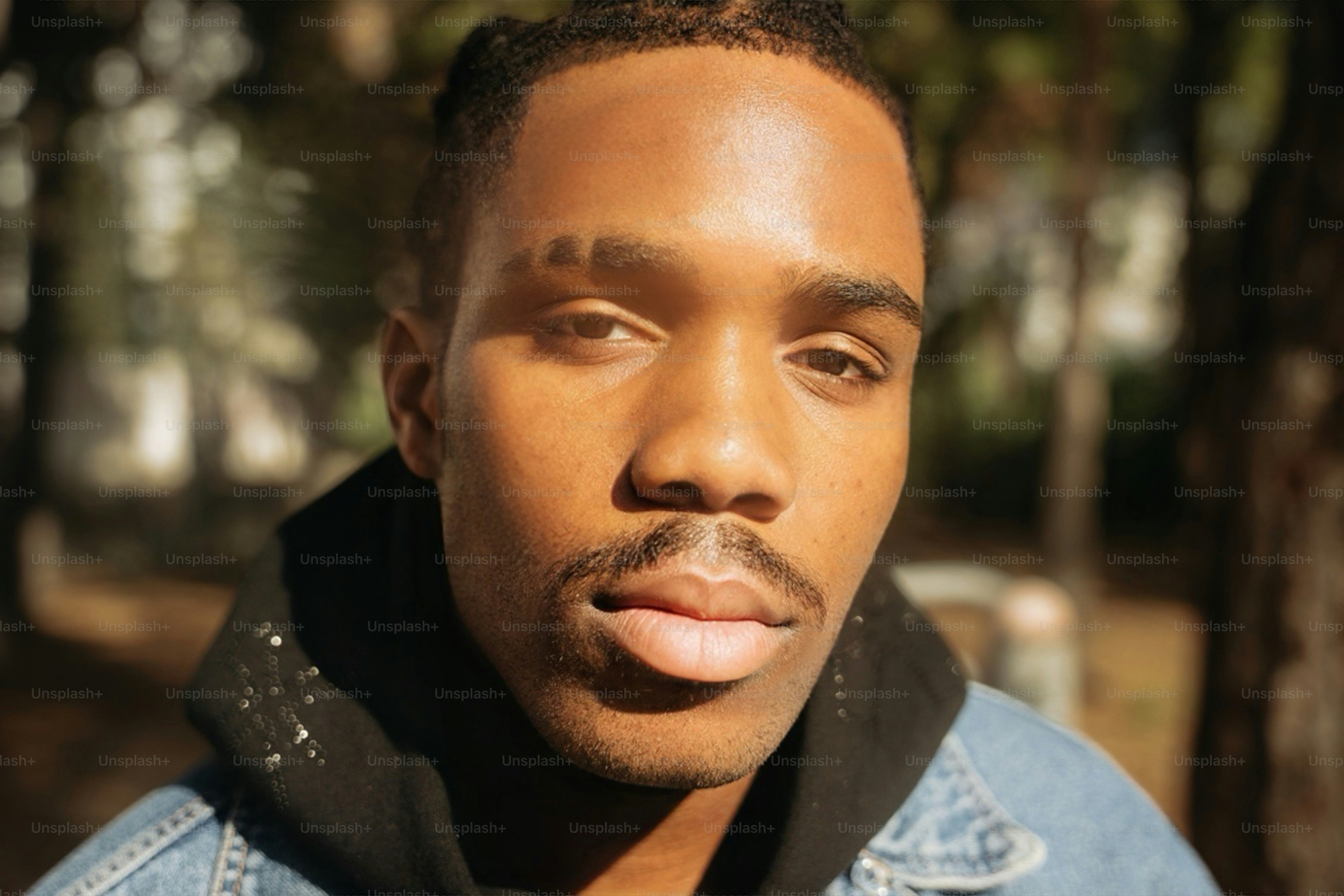 A man with a mustache and a shaved eyebrow line in a black hoodie and denim jacket