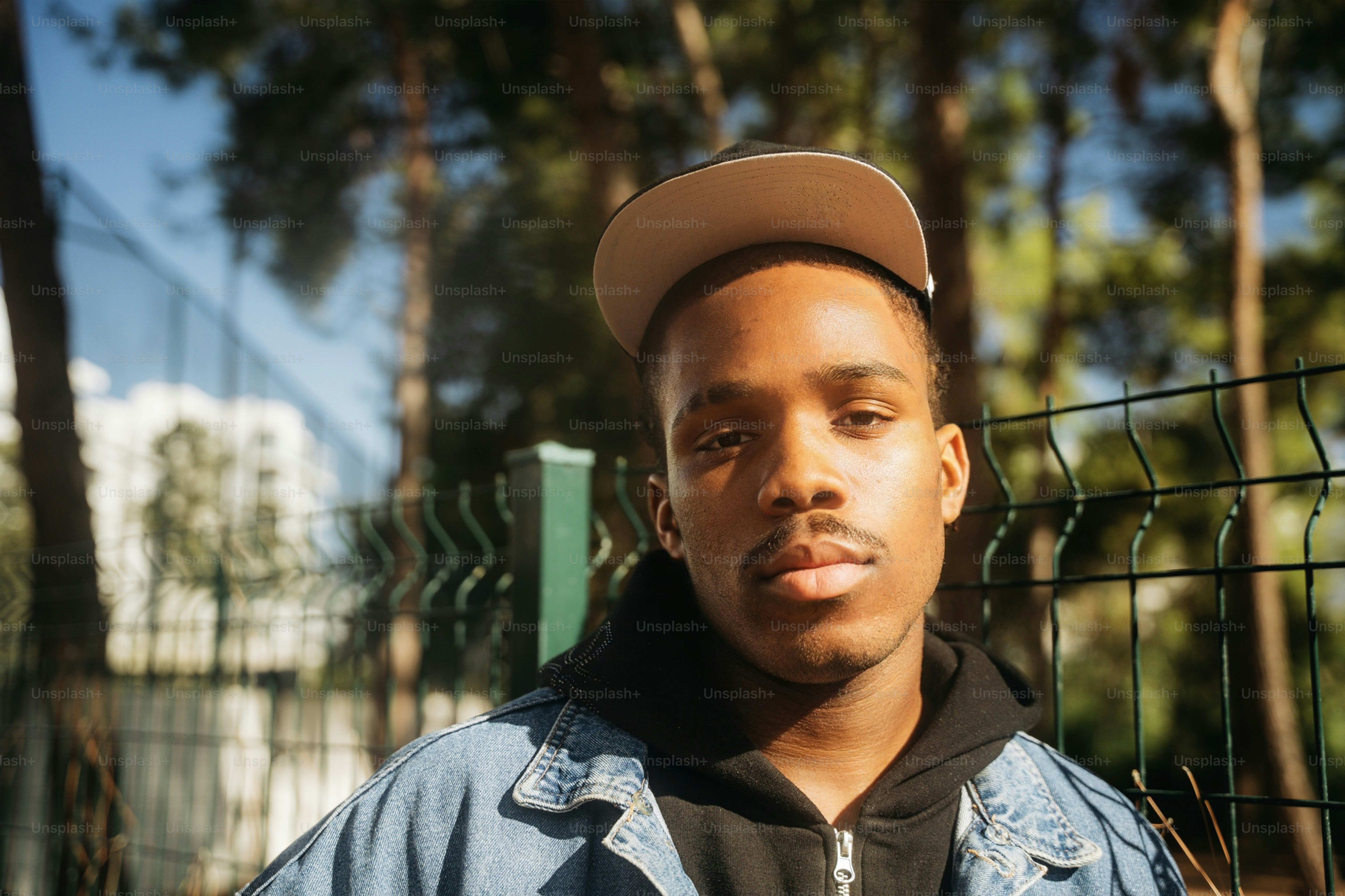 A young man in a denim jacket and baseball cap standing before a wire fence