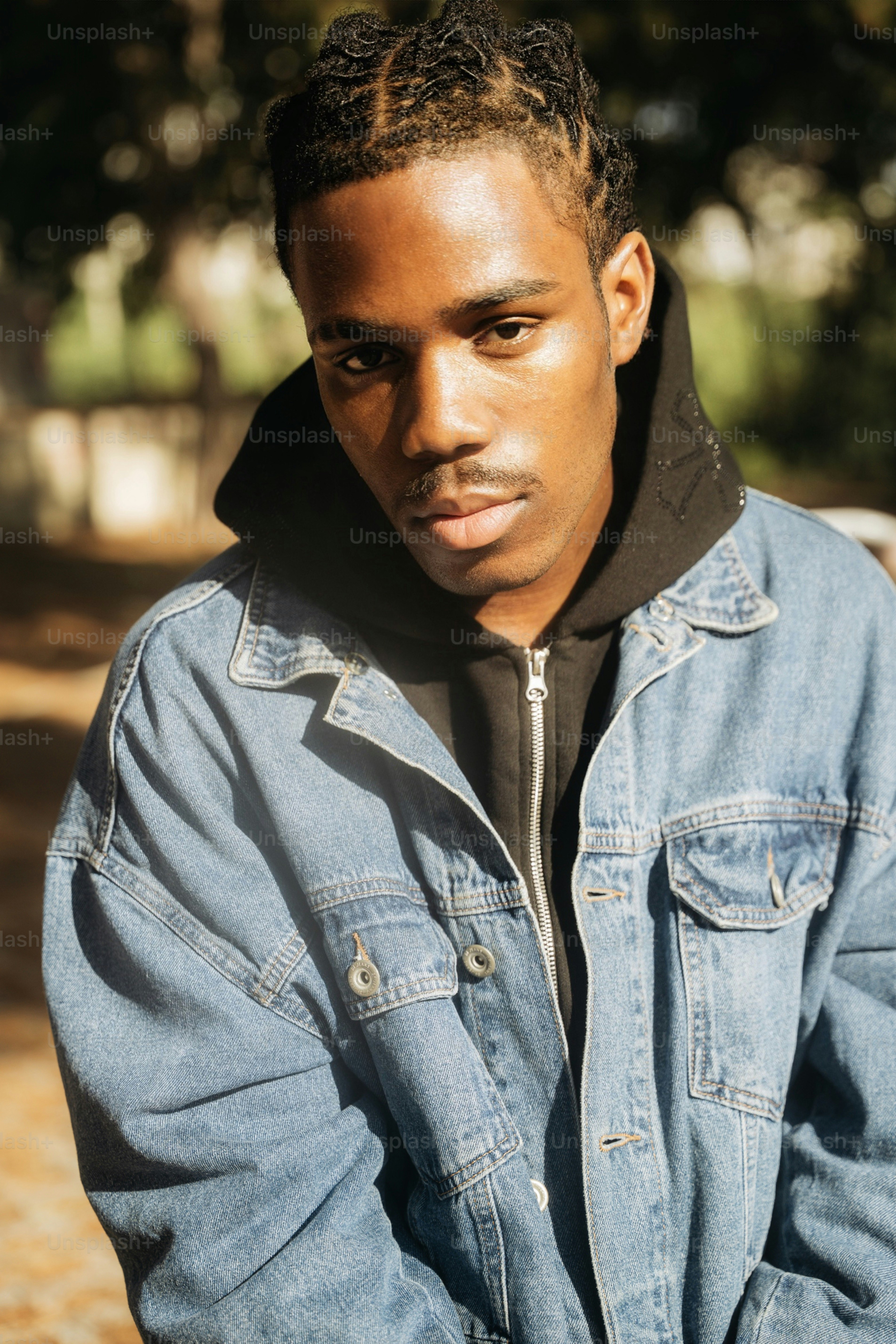 A young man with braided hair wearing a denim jacket and black hoodie in warm light