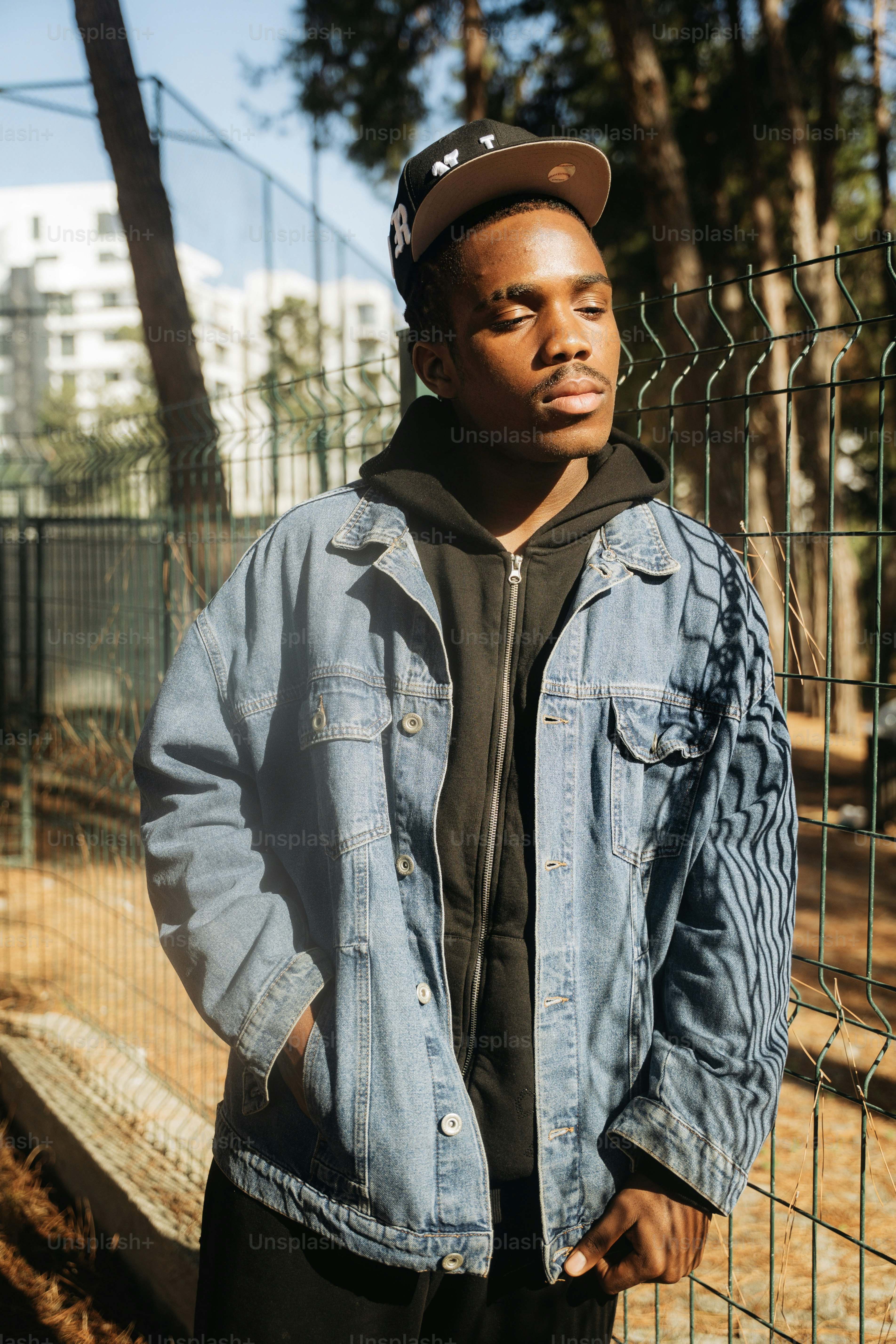 A young man in a denim jacket and black hoodie standing by a wire fence