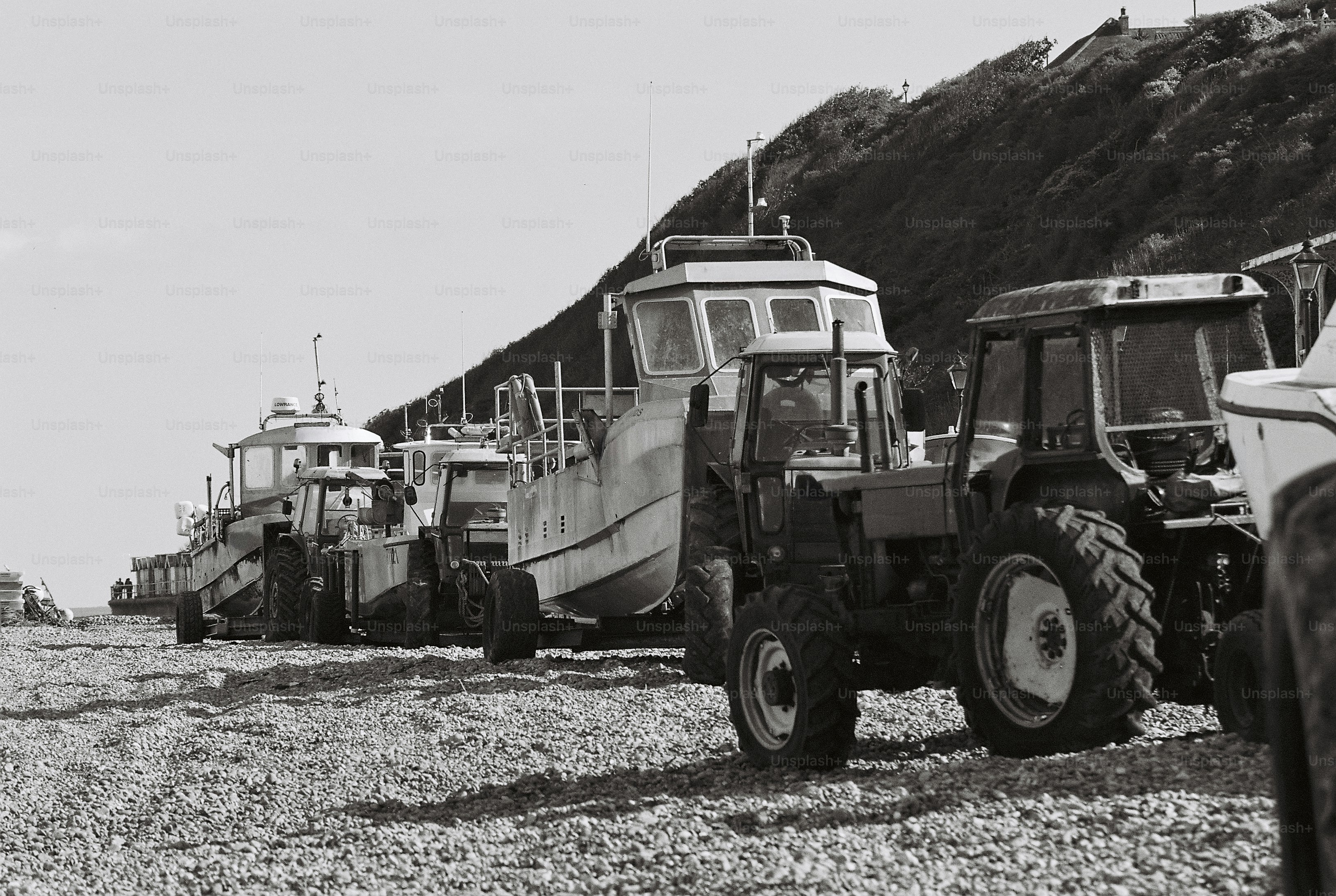 A line of fishing boats on trailers hitched to tractors on a pebble beach below a grassy cliff