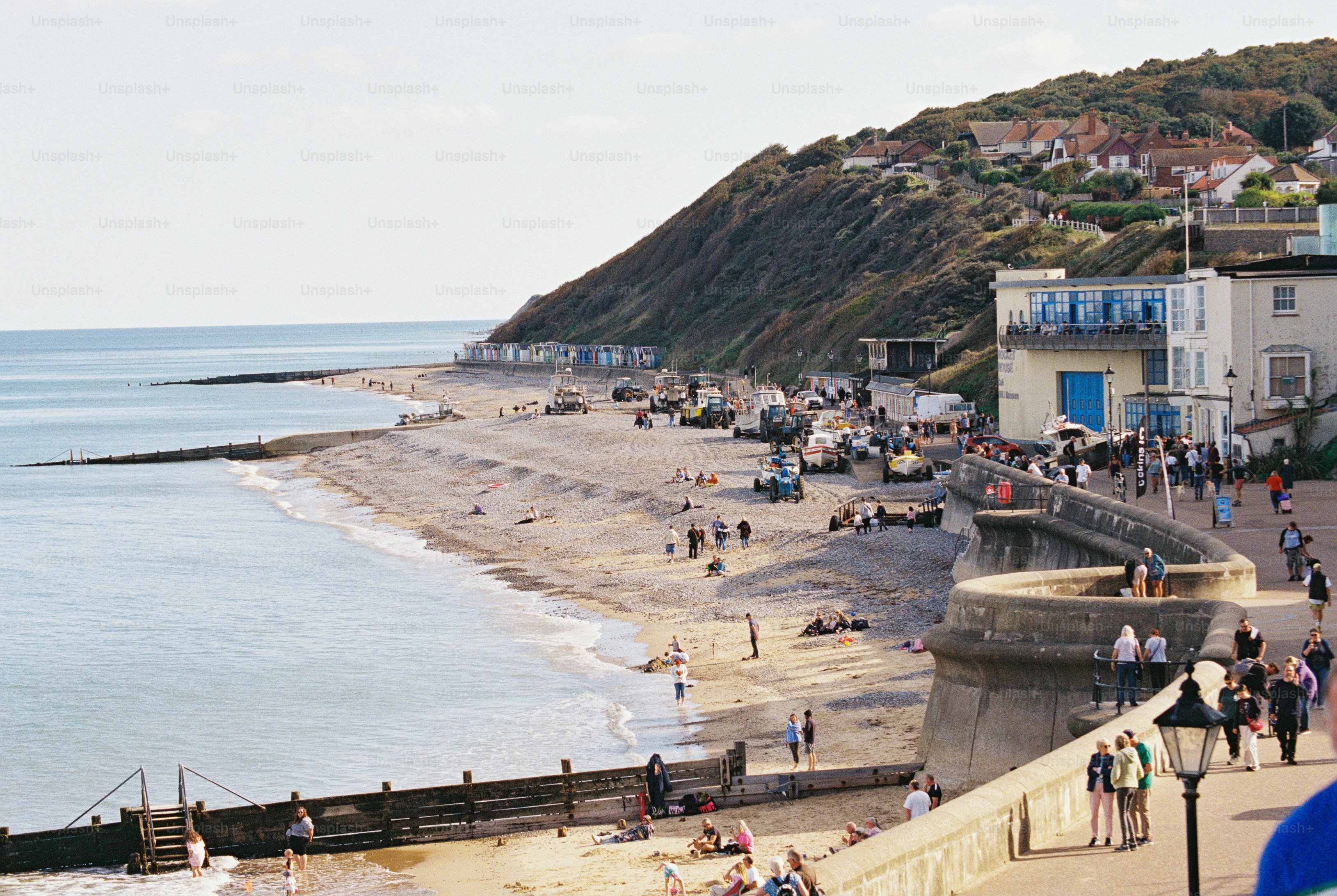 A curved sea wall and lamp post overlooking a pebble beach with boats and people below a cliff